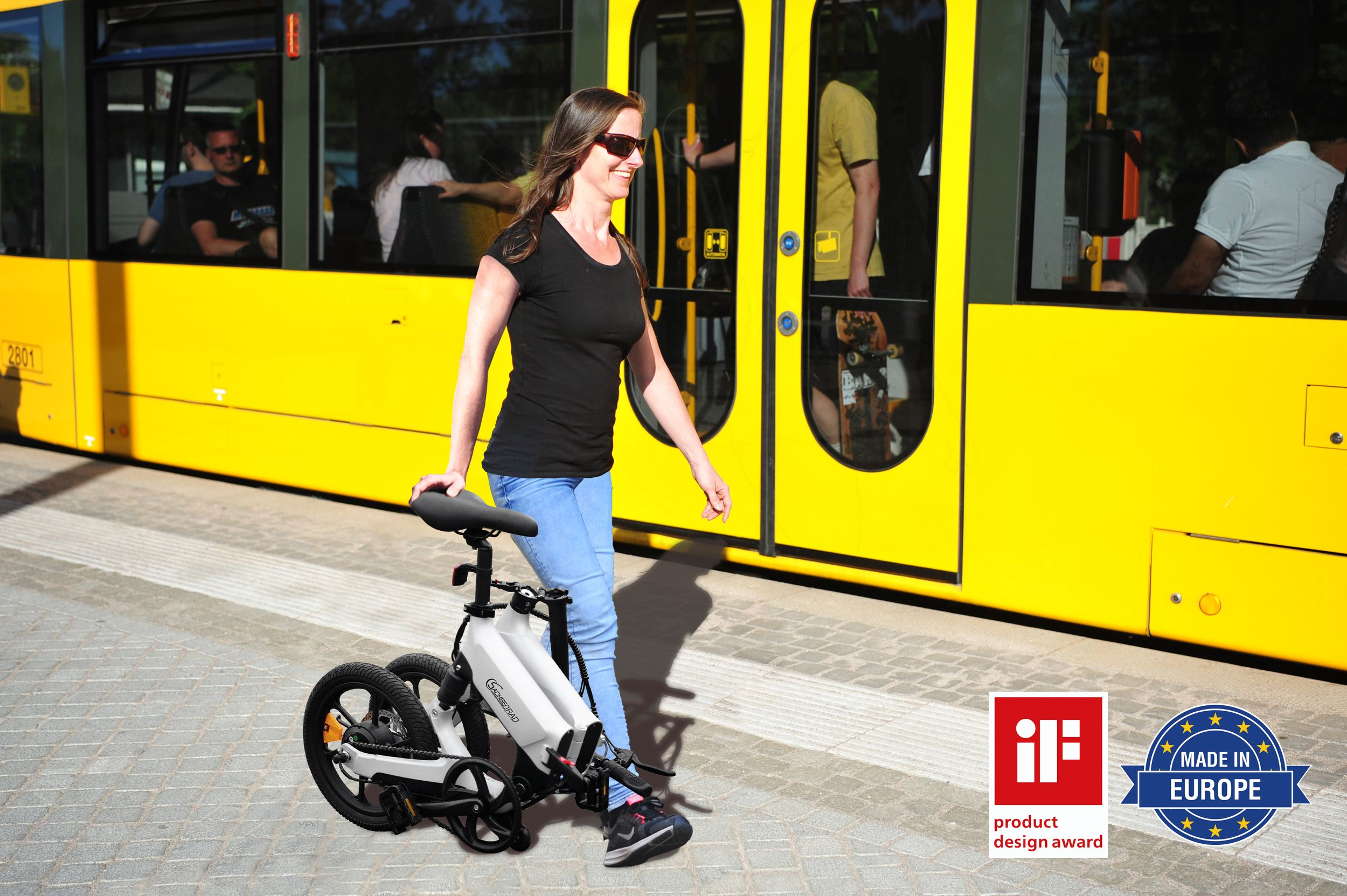Eine Frau geht mit einem weißen Faltrad neben einer gelben Straßenbahn.
