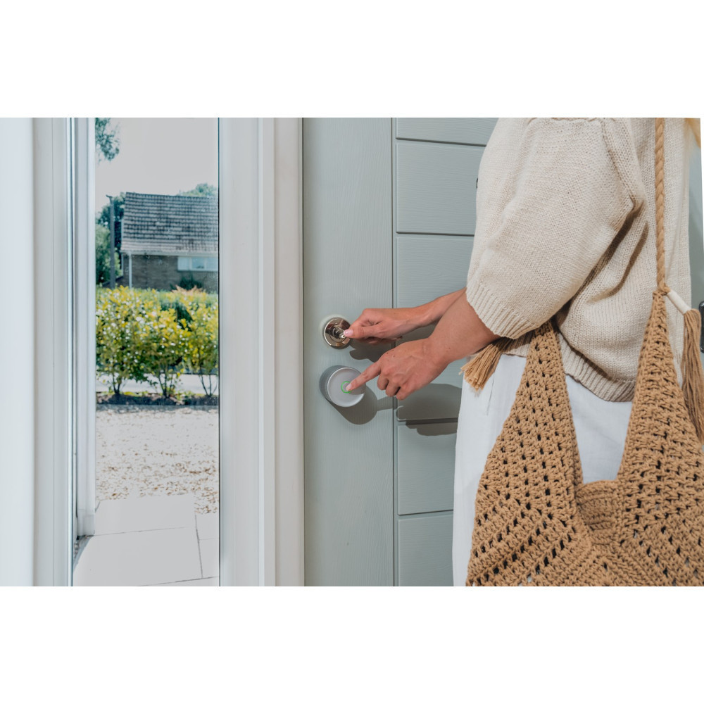 Eine Frau öffnet eine graue Tür mit einem Smart Lock, draußen ist ein Haus zu sehen.