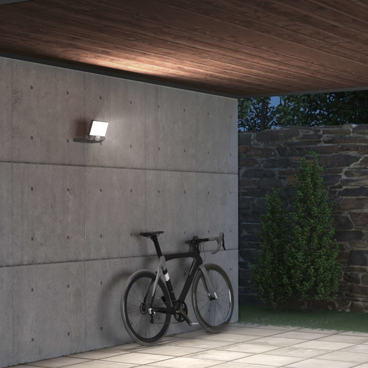 Ein schwarzes Rennrad lehnt an einer Betonwand unter einem Holzdach. Ein Licht ist an der Wand montiert.