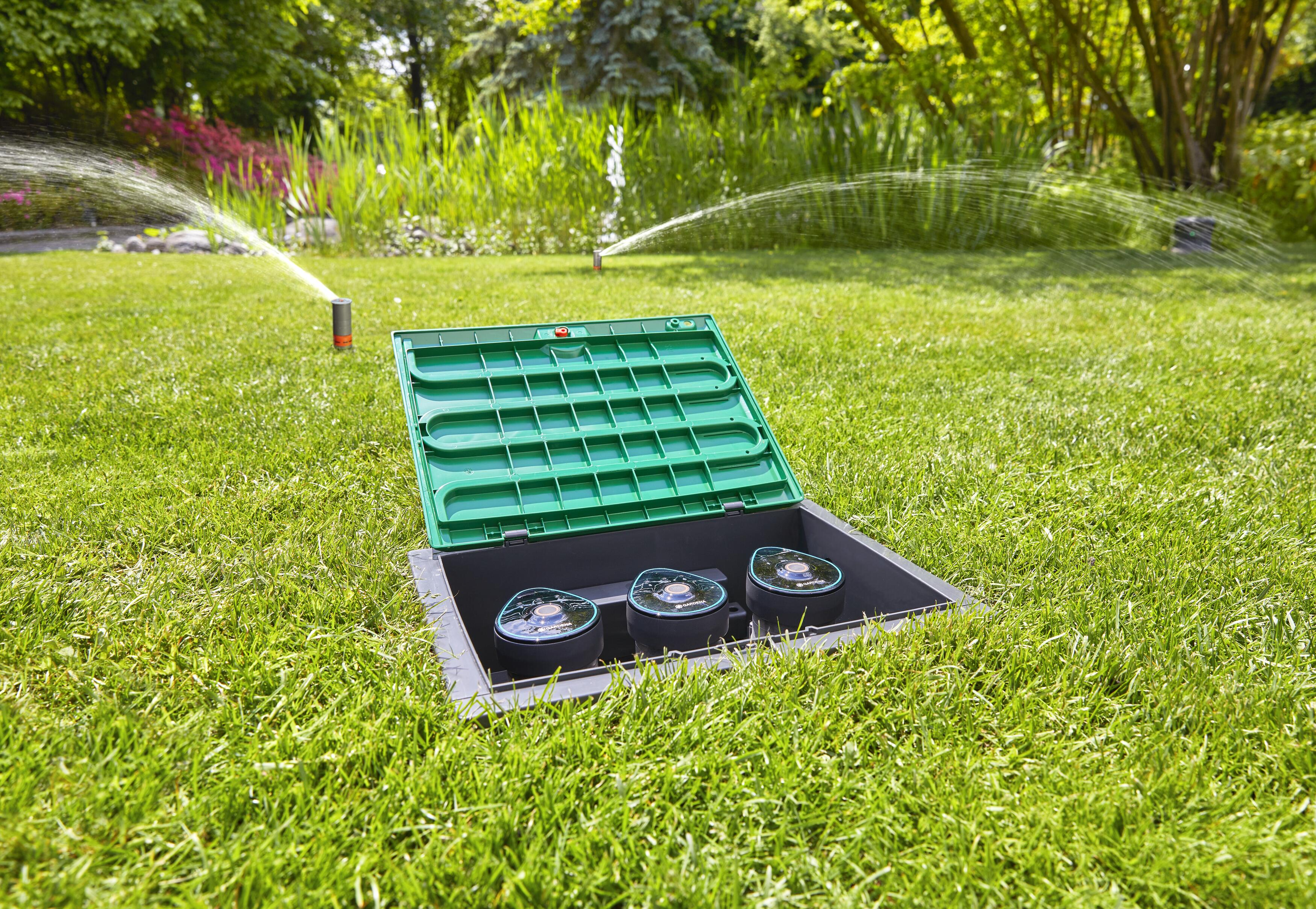 Ein offener grüner Bewässerungsautomatenkasten mit drei Sprinklern und einem Rasensprenger im Hintergrund.