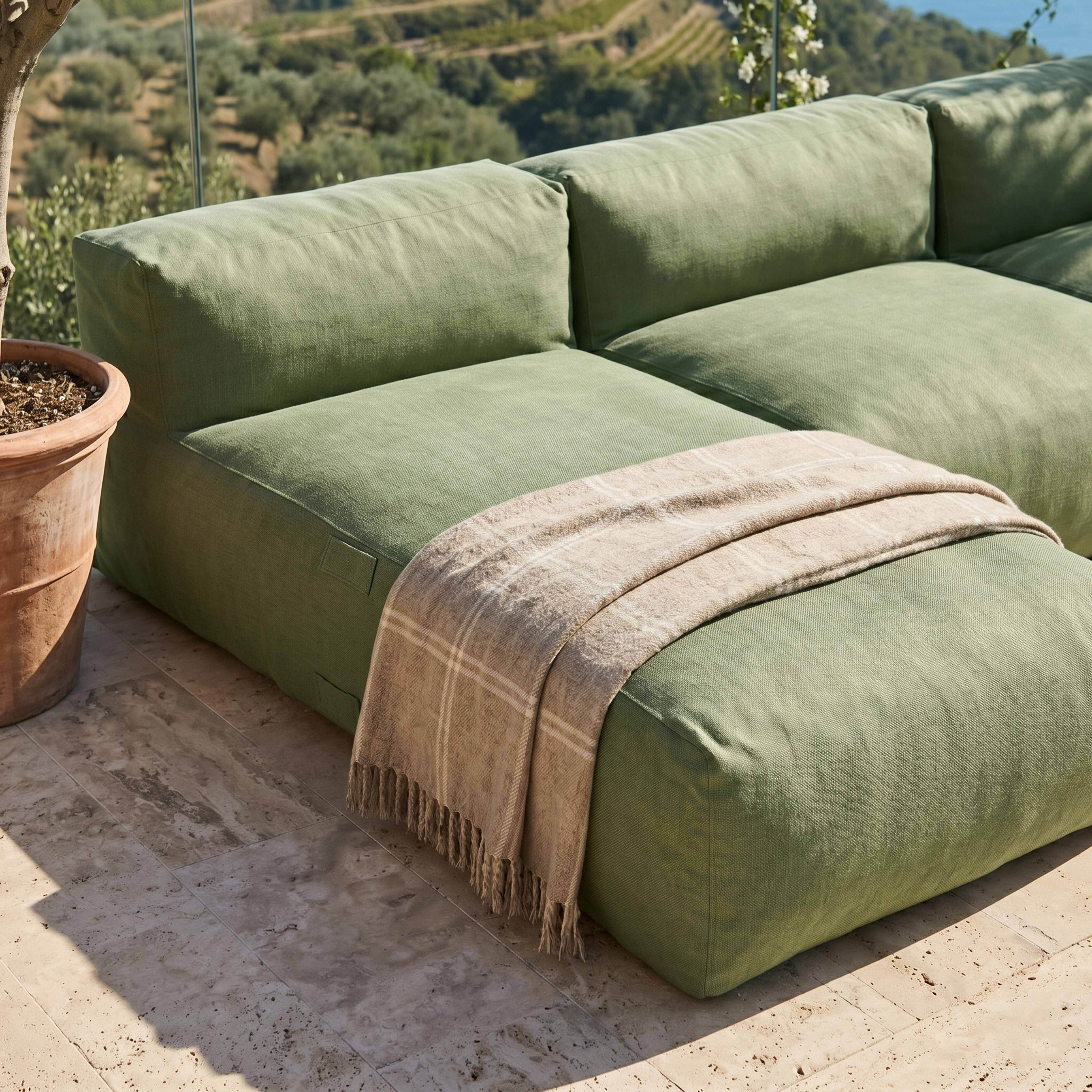 Grünes Outdoor-Sofa mit karierter Decke auf Steinboden, mit Bäumen und Meer im Hintergrund.