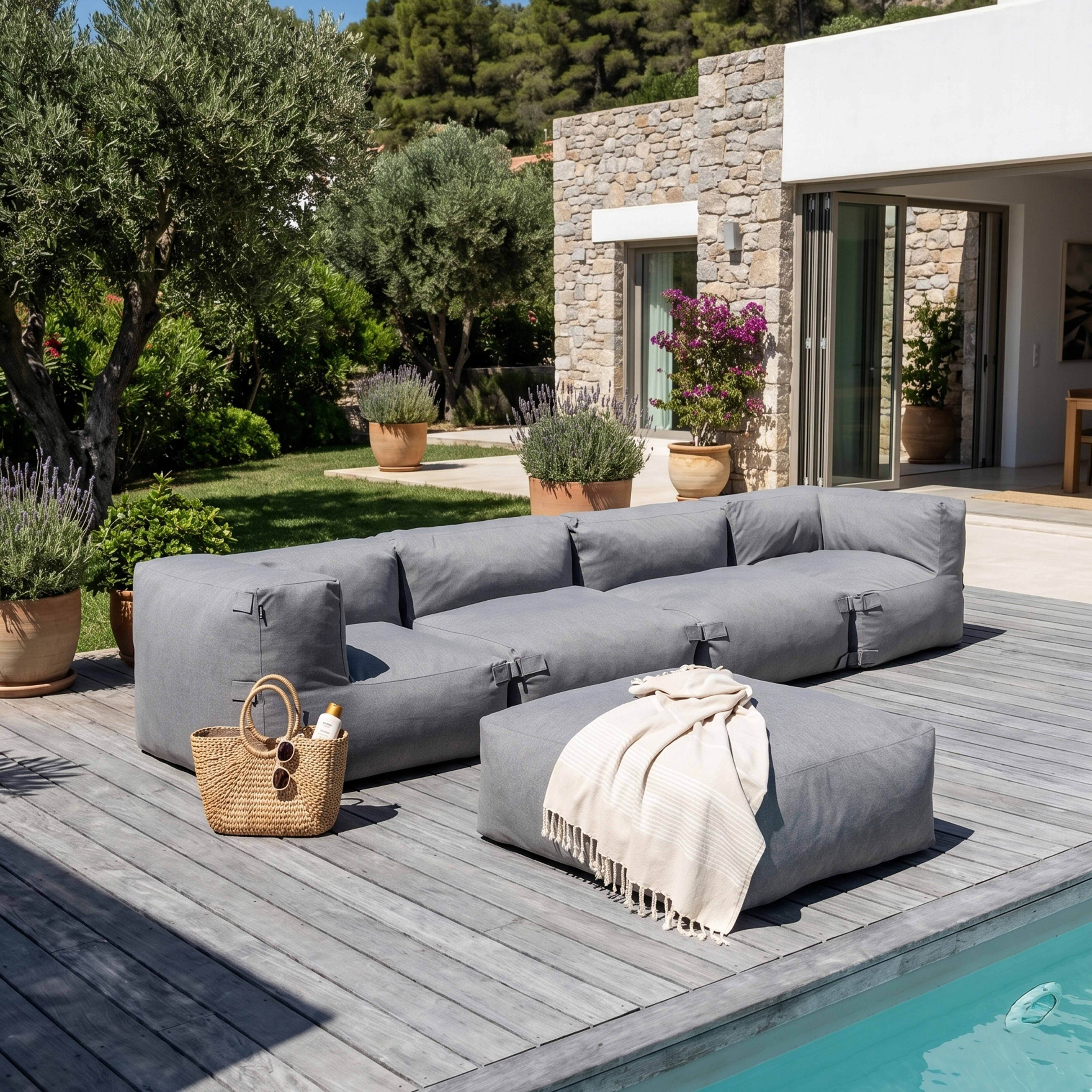Graues Outdoor-Sofa und Hocker auf Holzdeck neben Pool. Sonnencreme und Tasche.