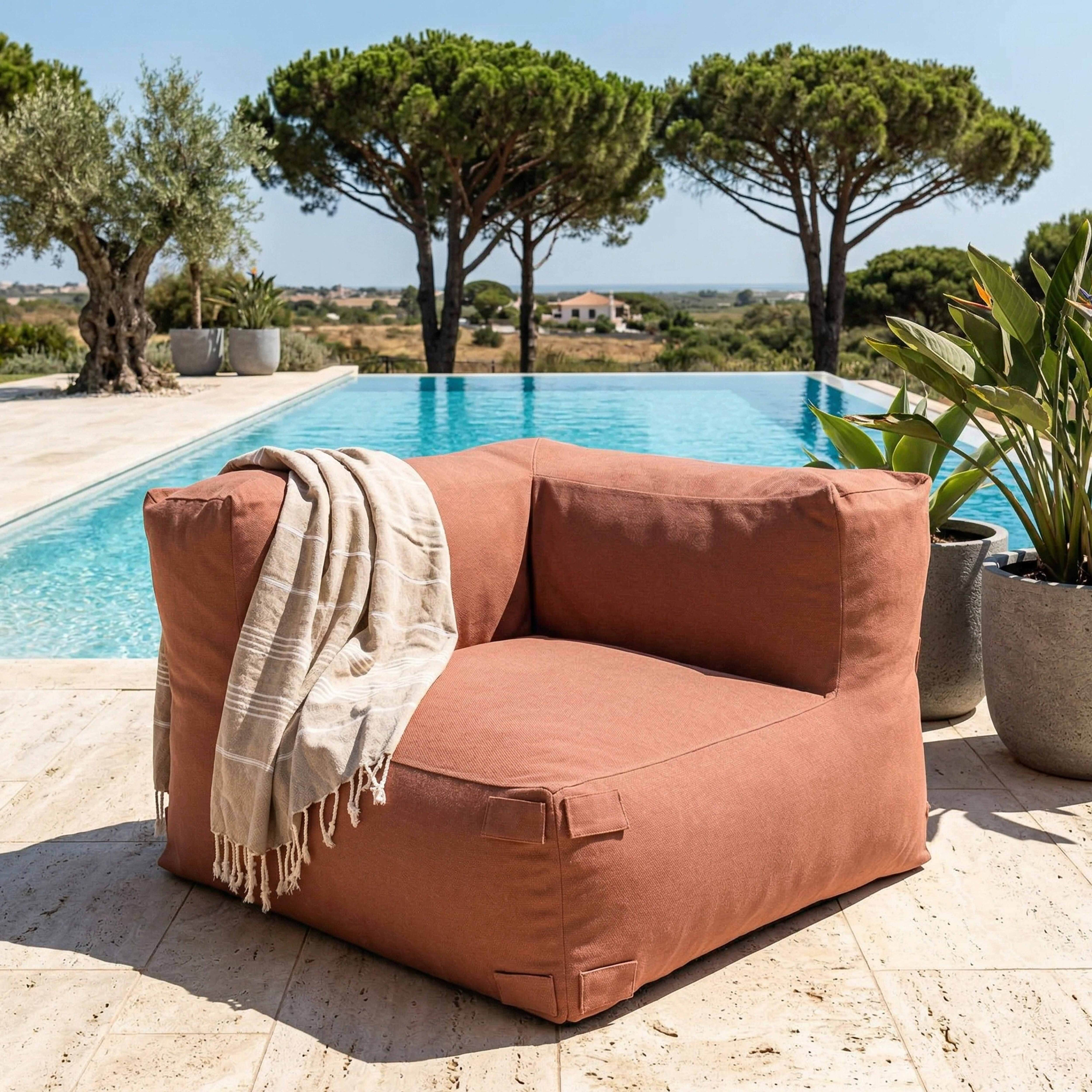 Ein korallenfarbener Sessel am Pool mit einer Decke, umgeben von Bäumen und Landschaft.