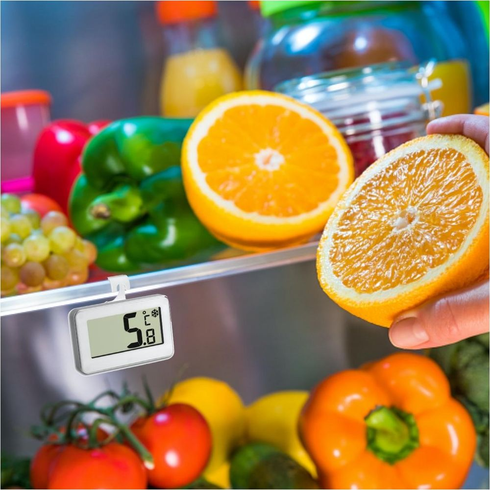 Ein Thermometer im Kühlschrank mit Früchten und Gemüse. Eine Hand hält eine Orangenhälfte.