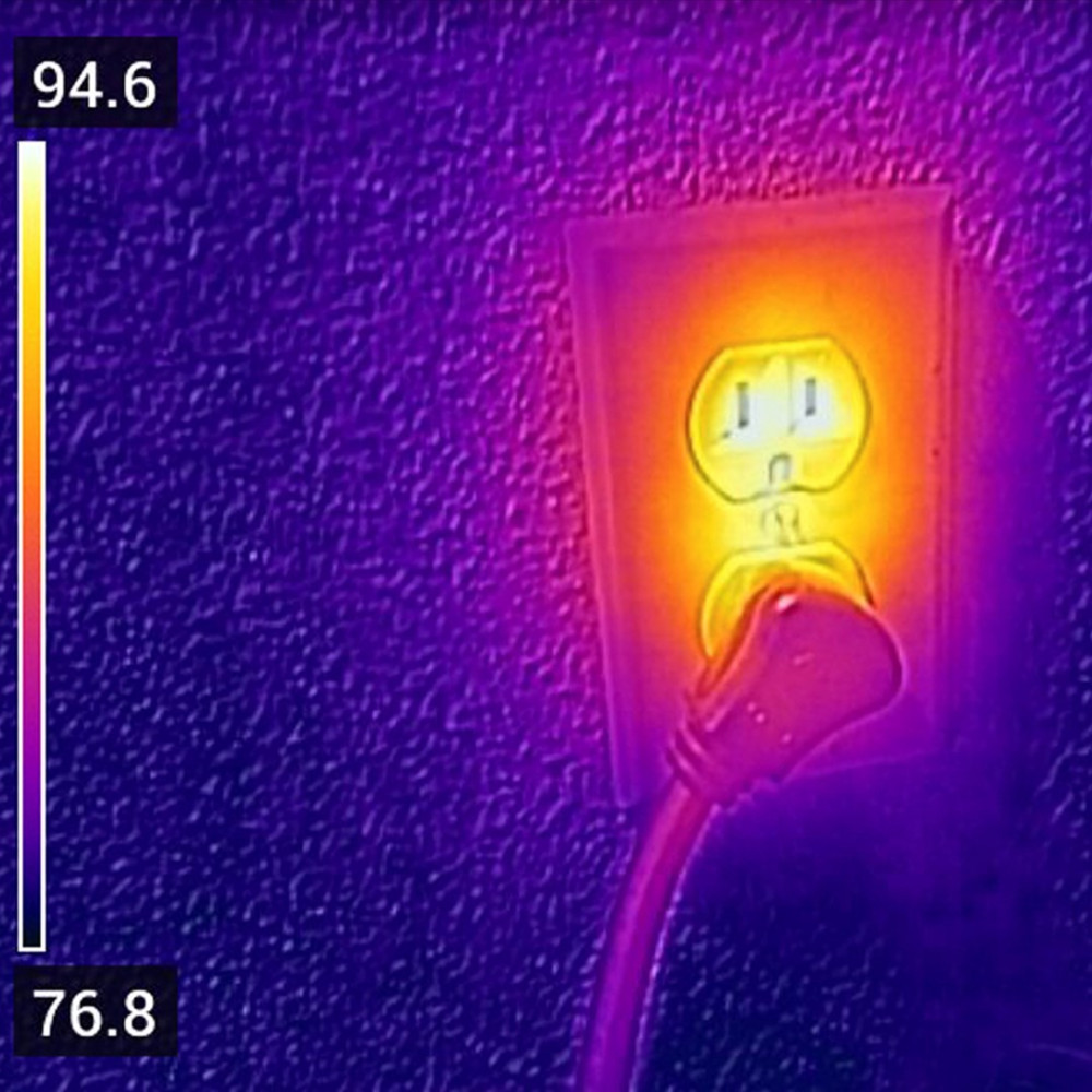 Obraz termowizyjny przedstawia gniazdko elektryczne z włożoną wtyczką, emitującą ciepło.