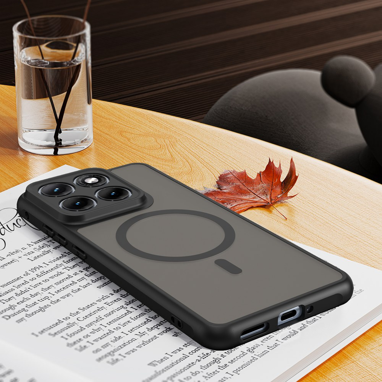 Een donkere telefoon met drie camera's ligt op een boek naast een glas water en een blad.