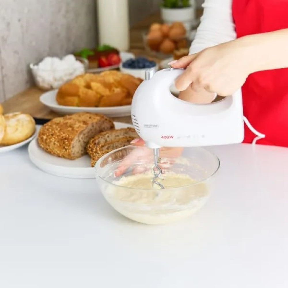 Ręka używa białego miksera ręcznego do mieszania ciasta w szklanej misce, z chlebem w tle.
