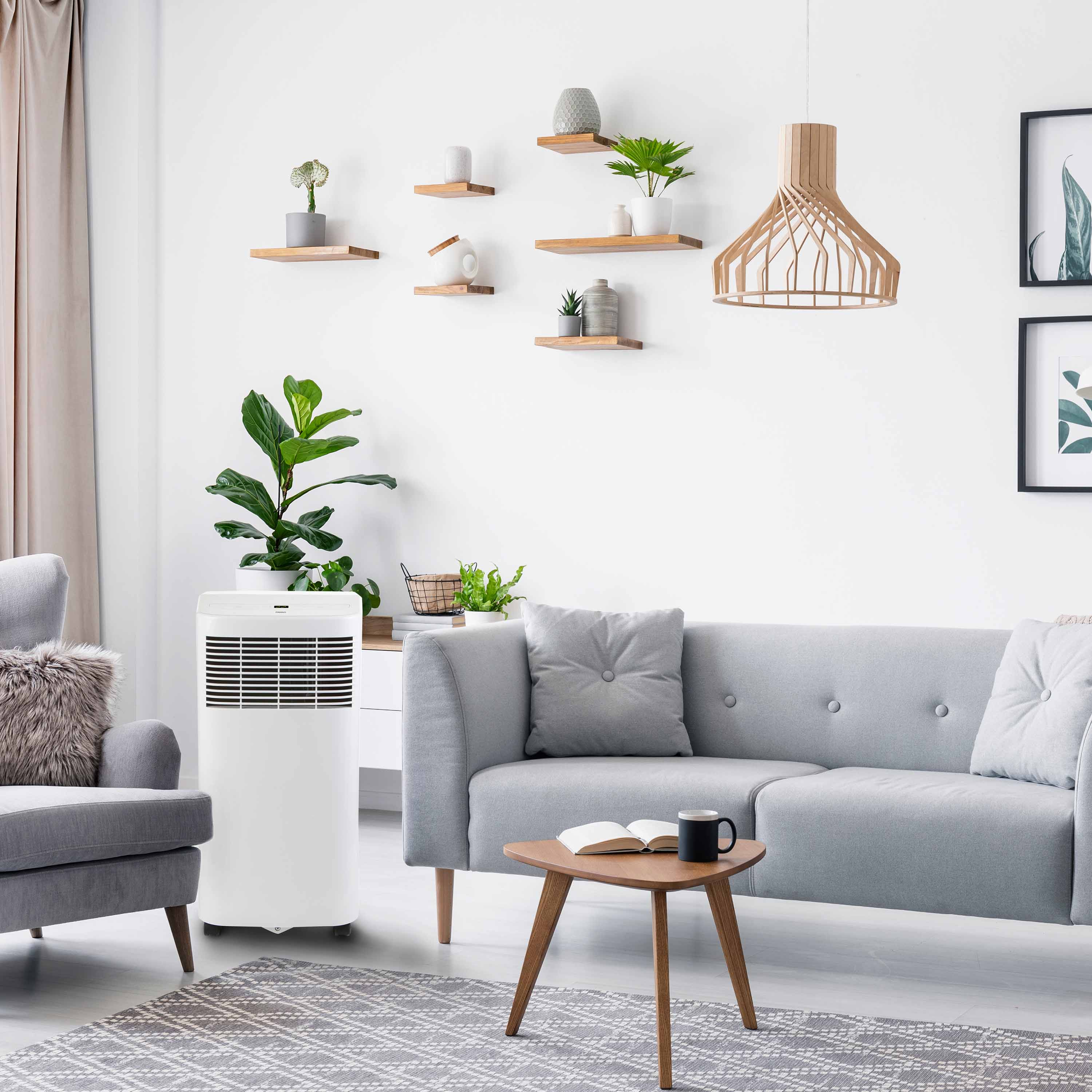 Een witte luchtkoeler naast een grijze fauteuil in een lichte woonkamer.