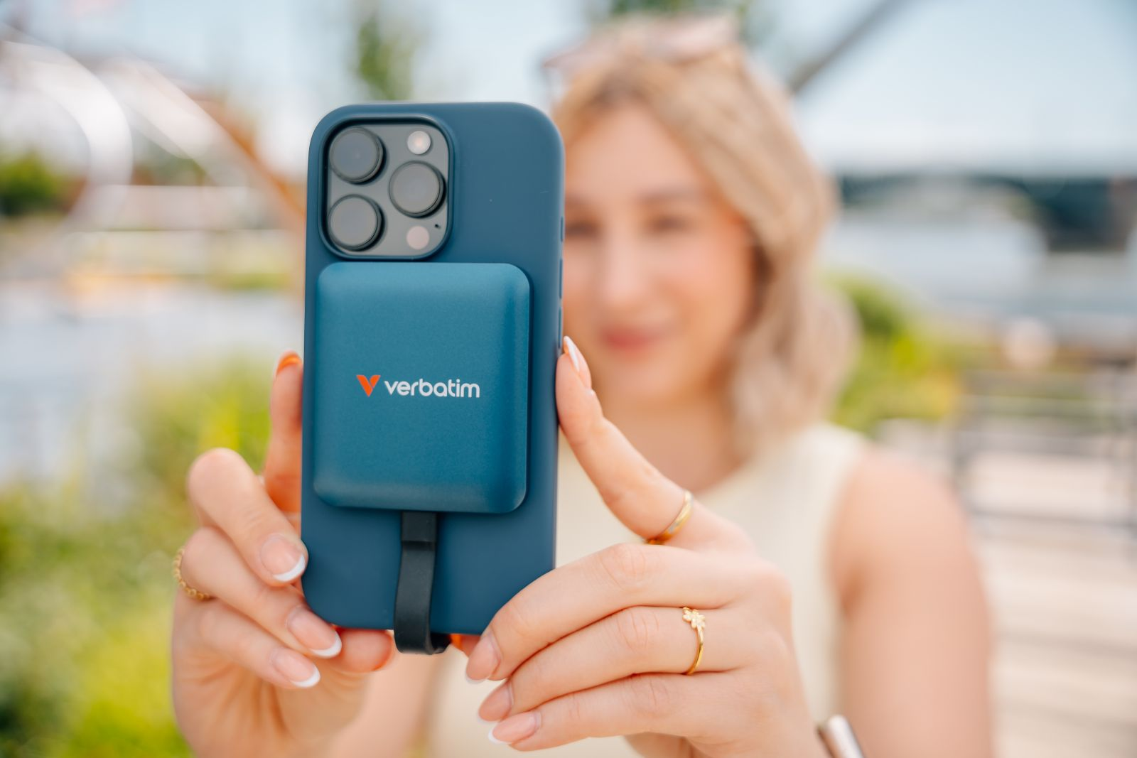 Eine Frau hält ein blaues Handy mit angeschlossenem Verbatim-Powerbank. Draußen.