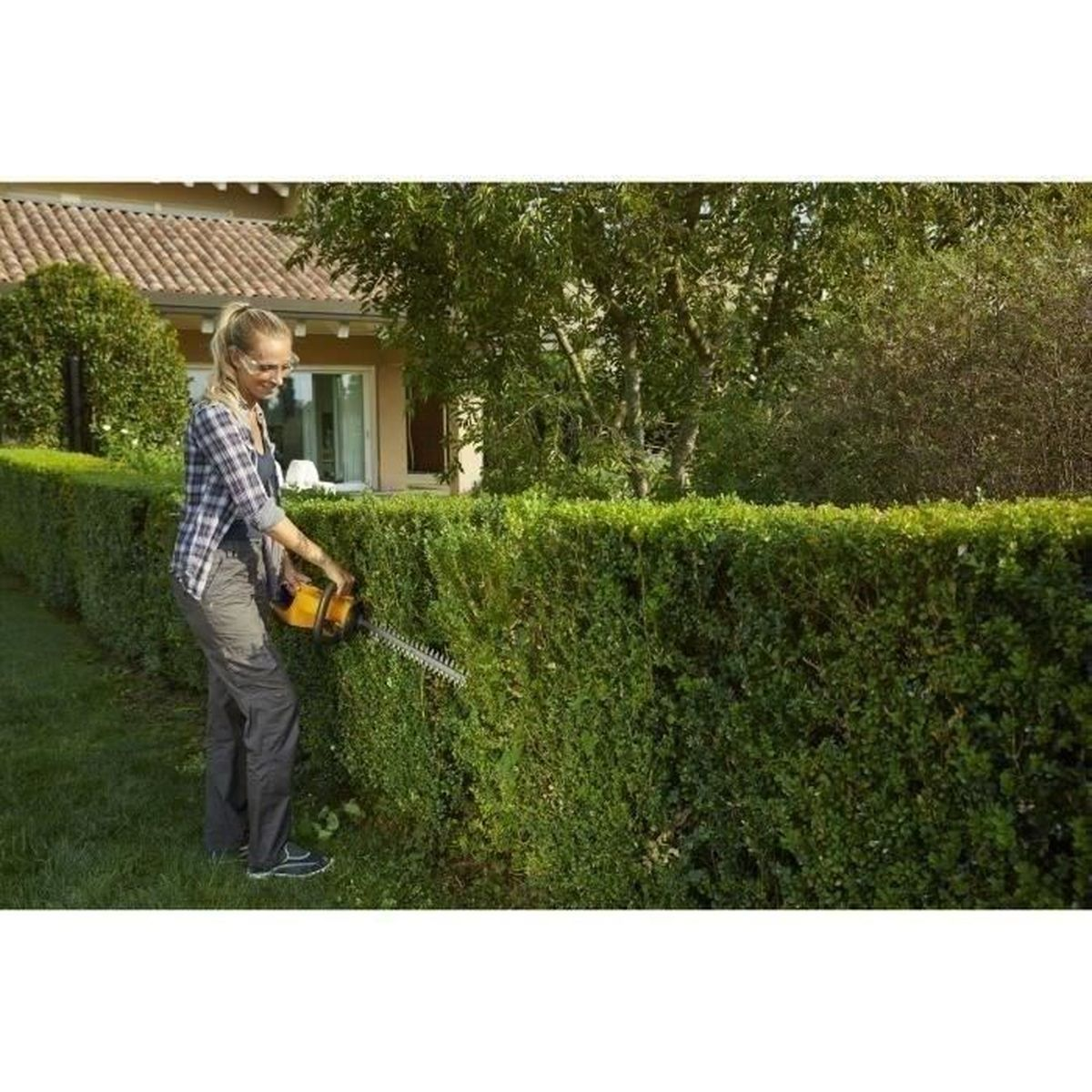 Frau trimmt Hecke mit elektrischem Heckenschneider in einem Garten.