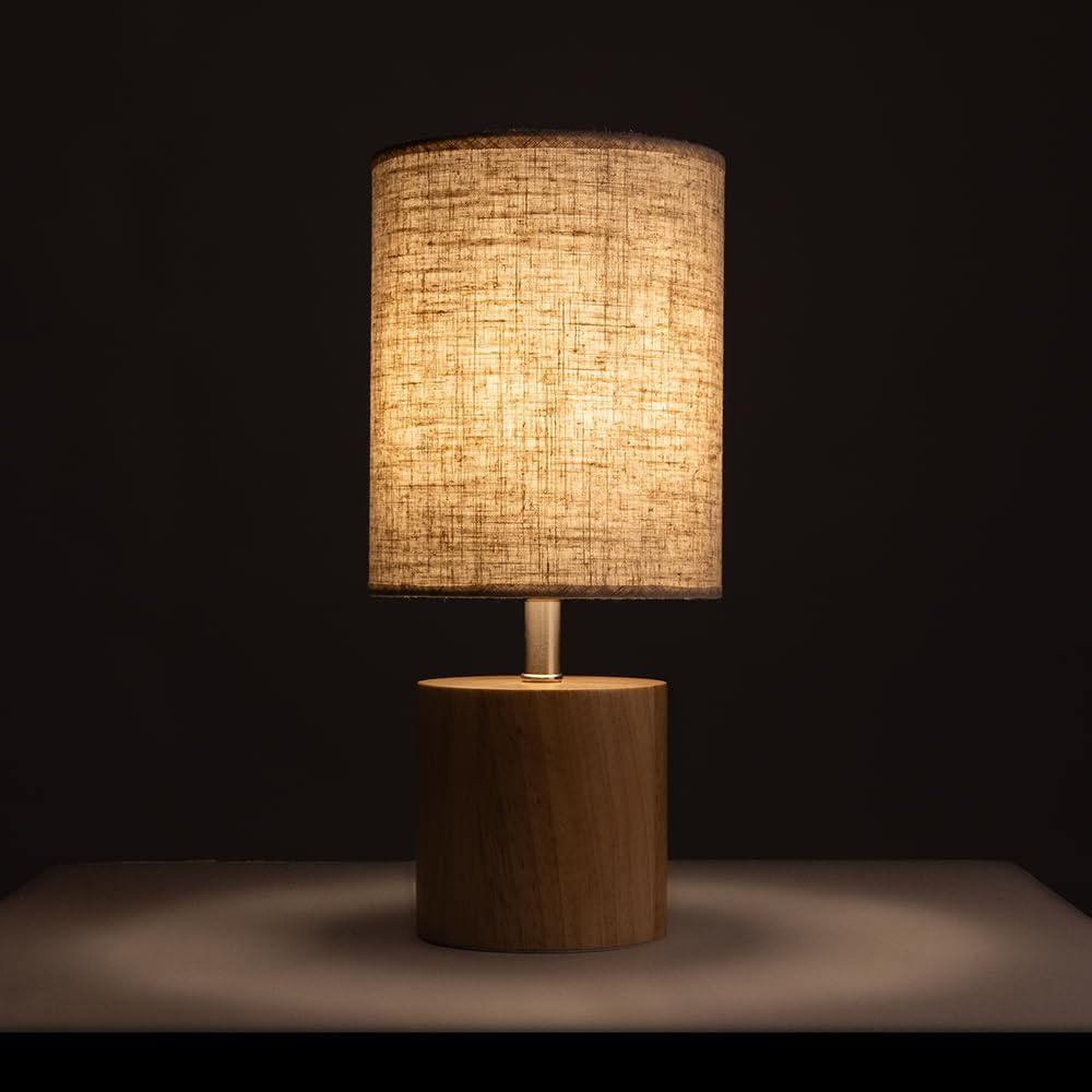 Eine beleuchtete Nachttischlampe mit Holzsockel und Leinenschirm. Dunkler Hintergrund.