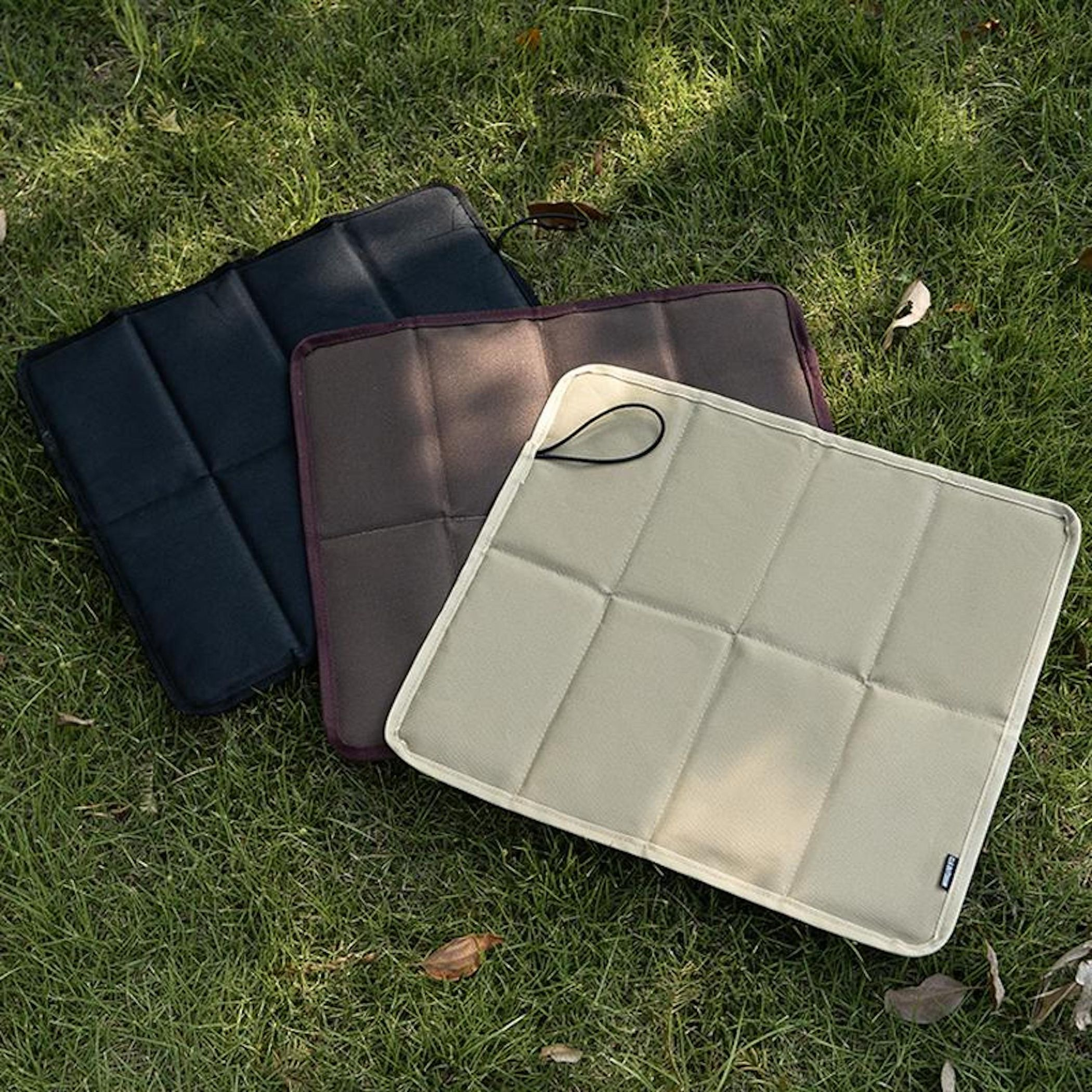 Drei gefaltete Campingmatten in Schwarz, Braun und Beige liegen auf grünem Gras.