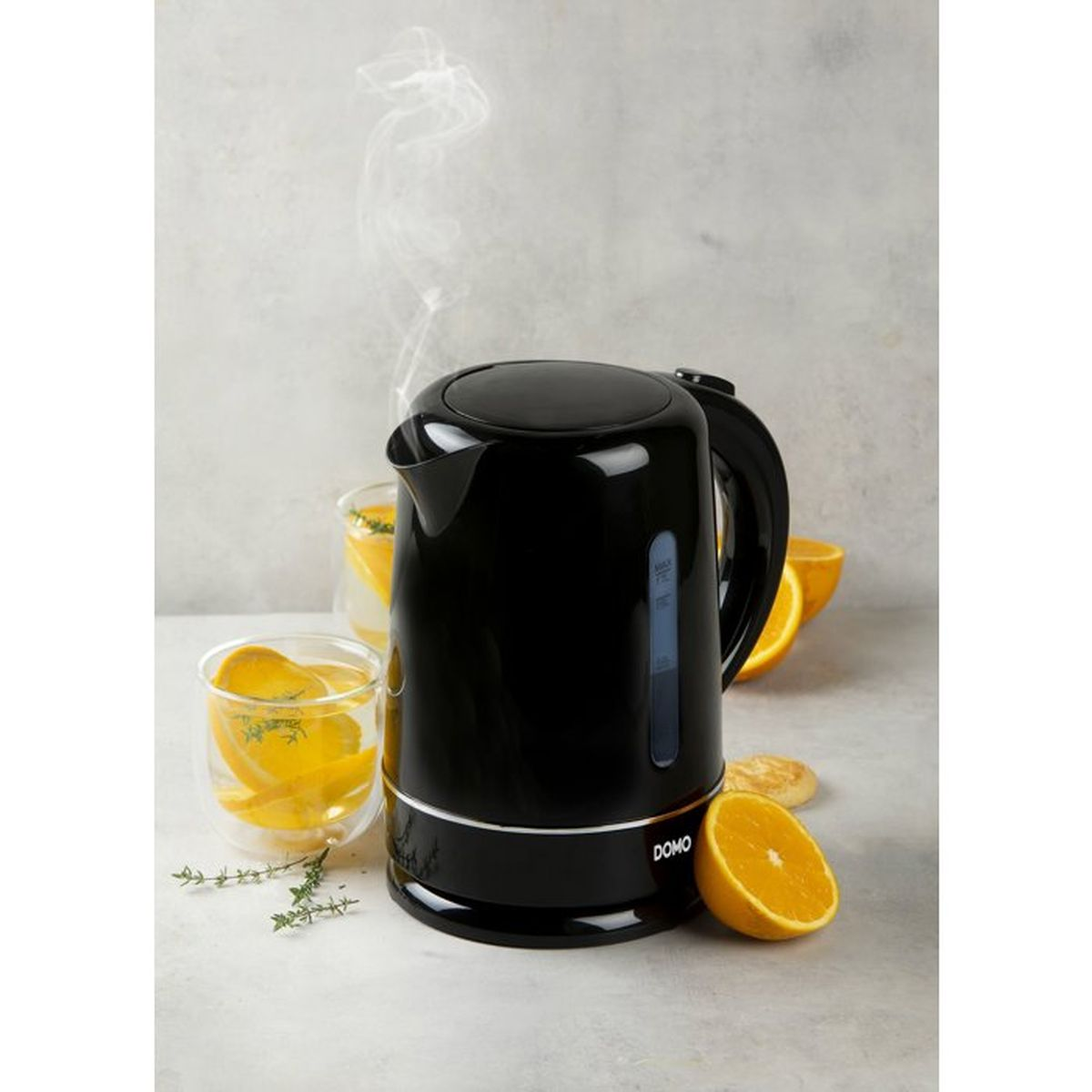 Schwarzer Wasserkocher mit dampfendem Wasser, daneben Gläser mit Orangen und Kräutern.