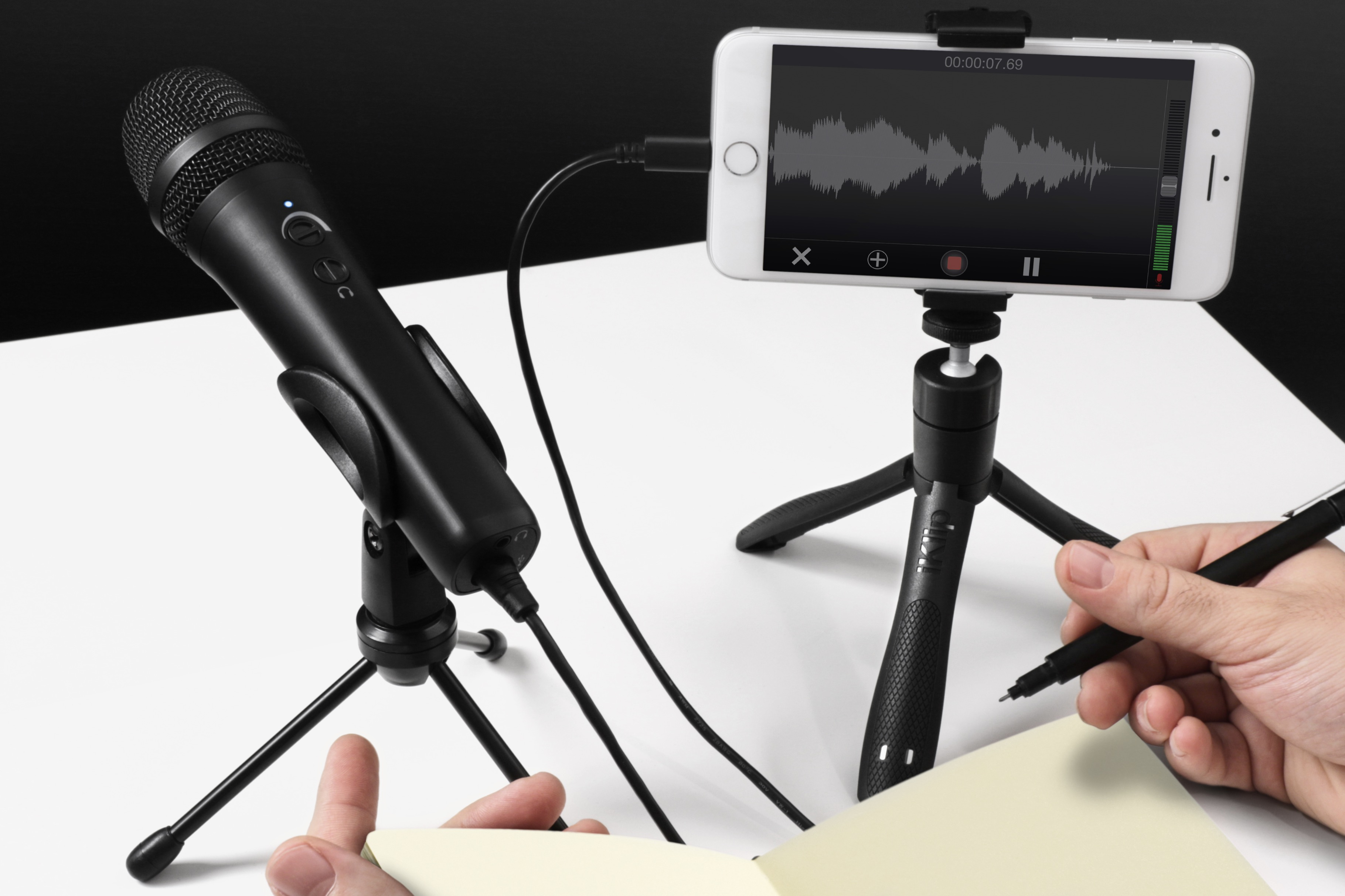 Schwarzes Mikrofon, Smartphone mit Wellenform-Anzeige, Stift und Notizbuch auf weißem Tisch.