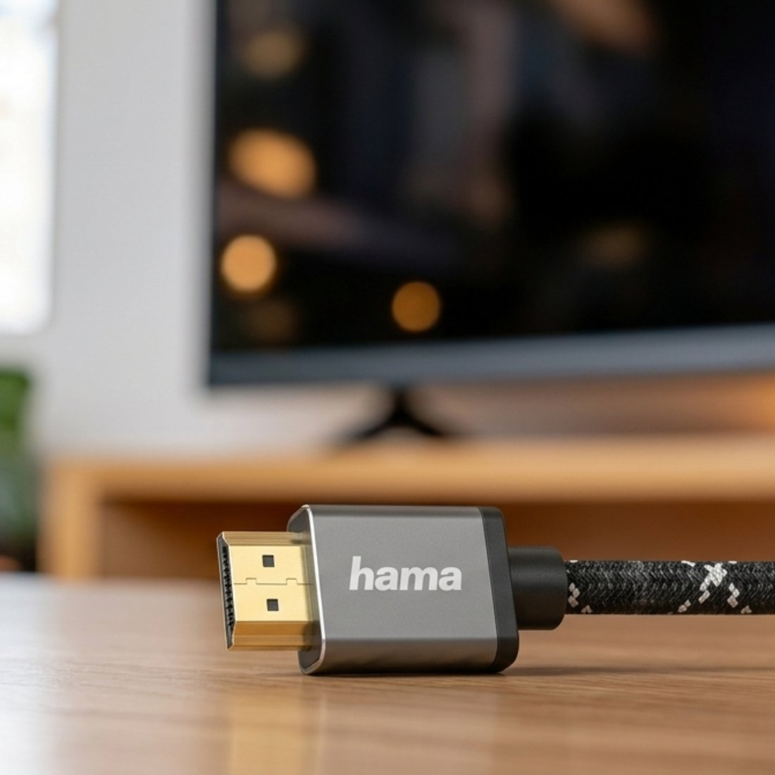 Kabel HDMI Hama ze złotą wtyczką leży na drewnianym stole, w tle telewizor.