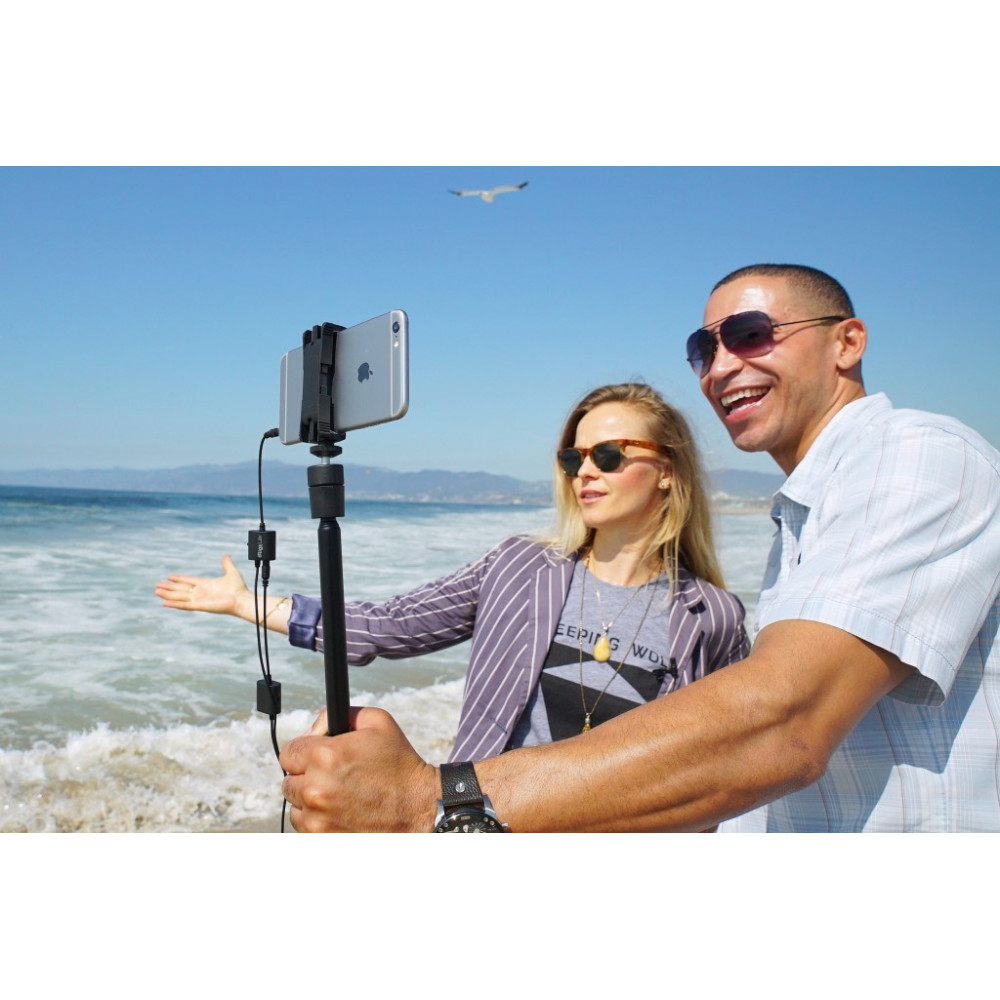 Para na plaży filmuje telefonem na selfie sticku, w tle ocean i niebo.
