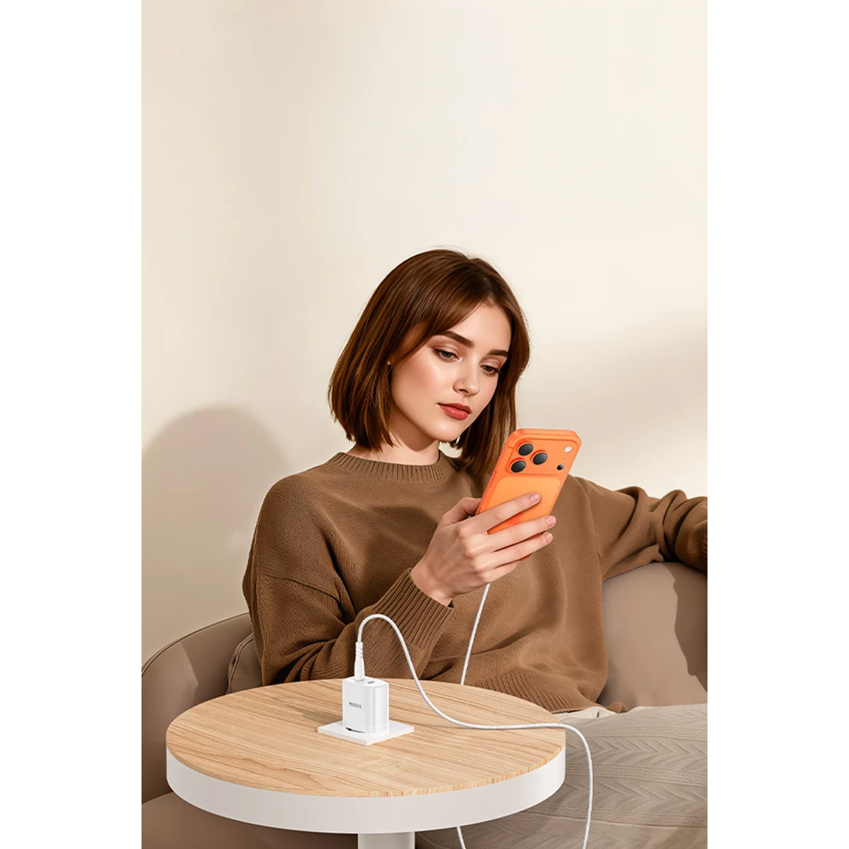 Kahverengi kazaklı kadın, ahşap bir masada beyaz şarj cihazıyla turuncu telefonunu şarj ediyor.