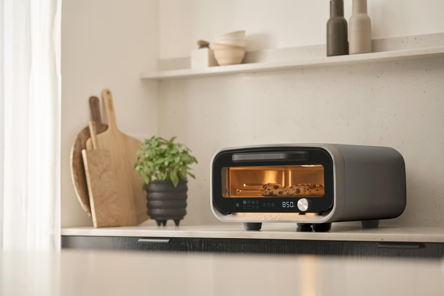 Un piccolo forno su un bancone con una pizza all'interno. Taglieri di legno e una pianta accanto.