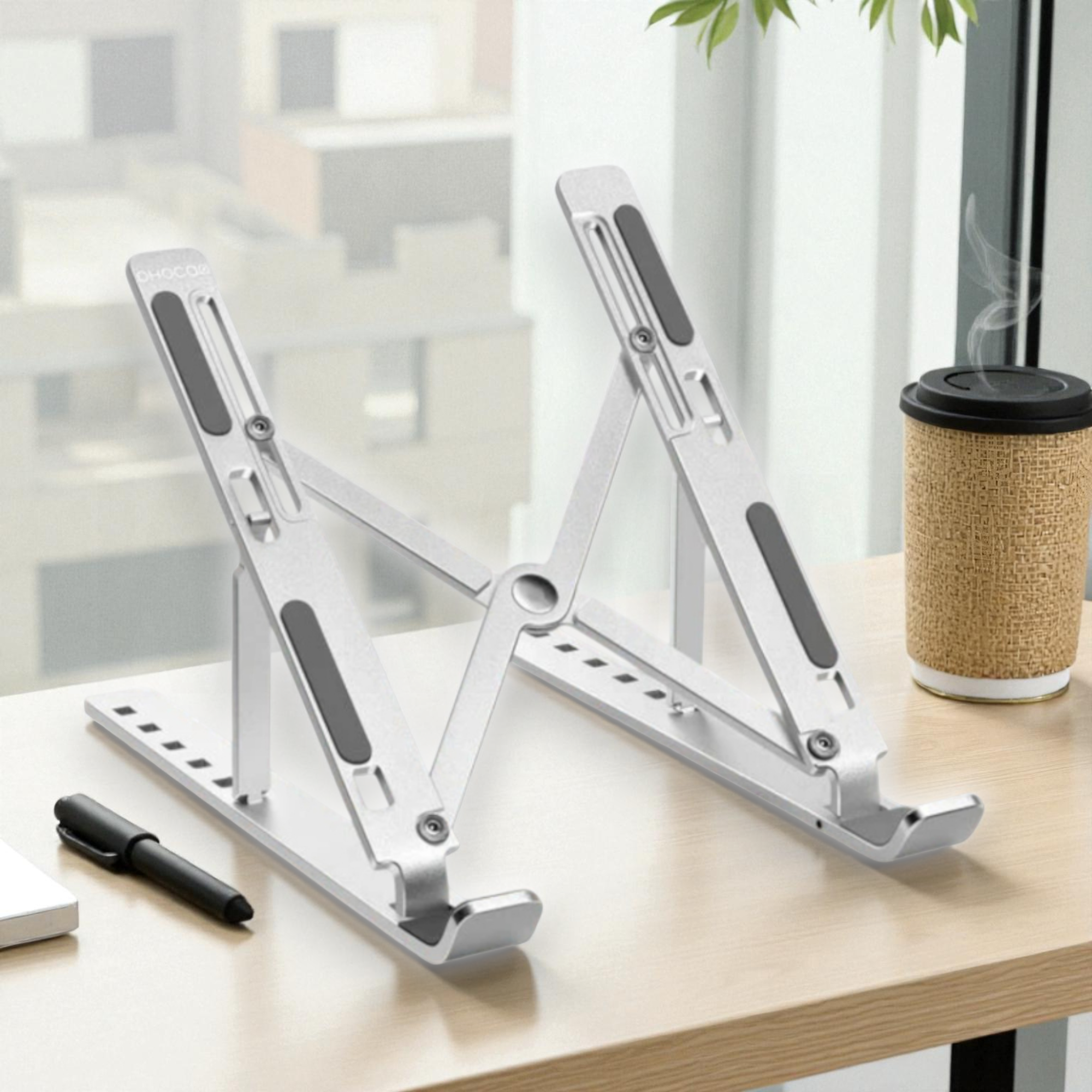 Soporte plateado ajustable para portátil sobre escritorio de madera con bolígrafo y taza.