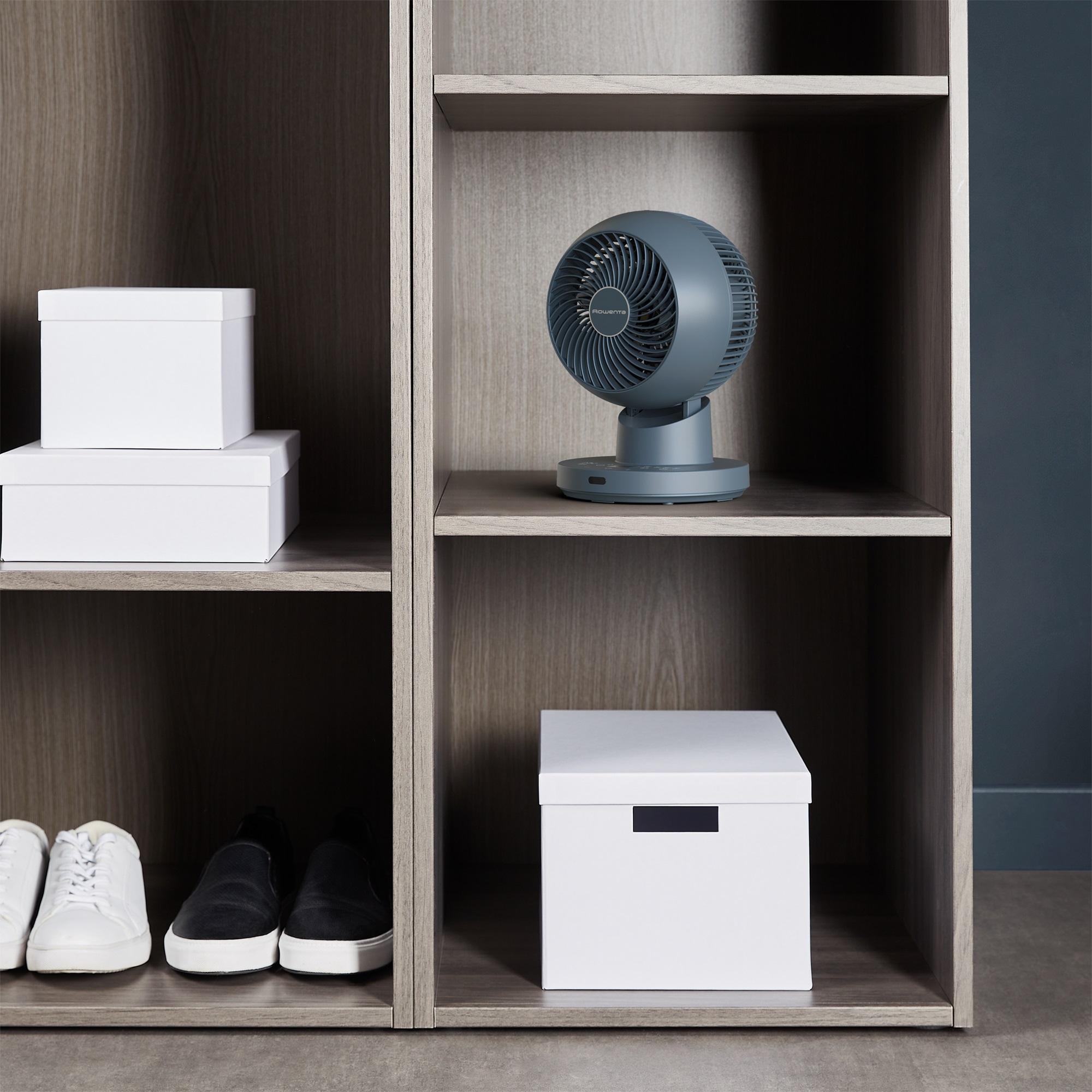 Ein grauer Ventilator auf einem Regal über einer weißen Box. Zwei weiße Boxen sind daneben.