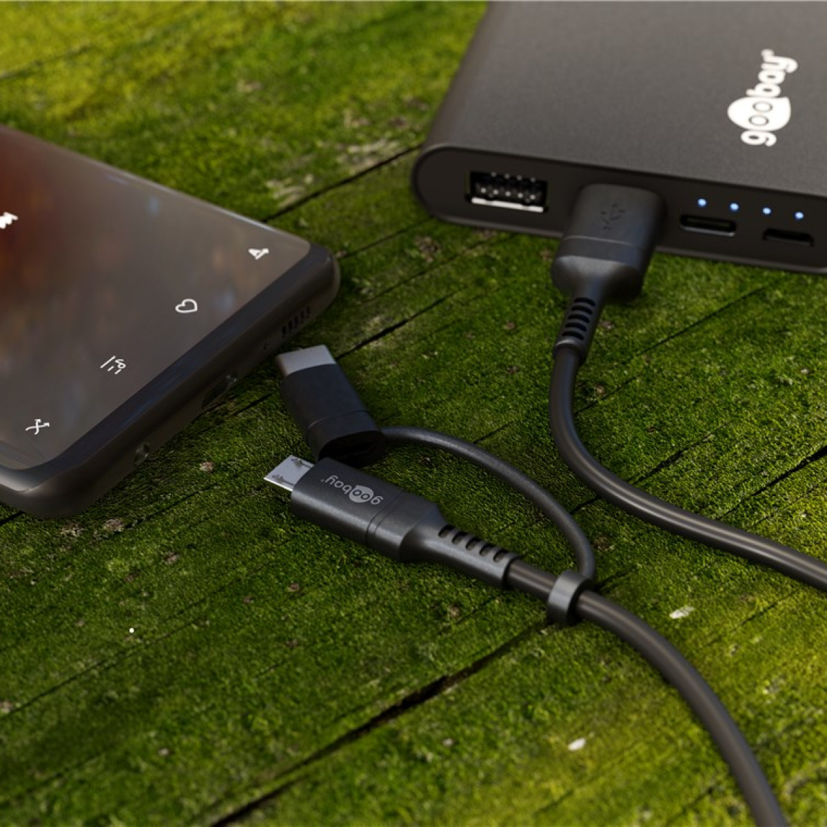 Uno smartphone e un power bank neri collegati a un cavo USB su terreno muschioso.