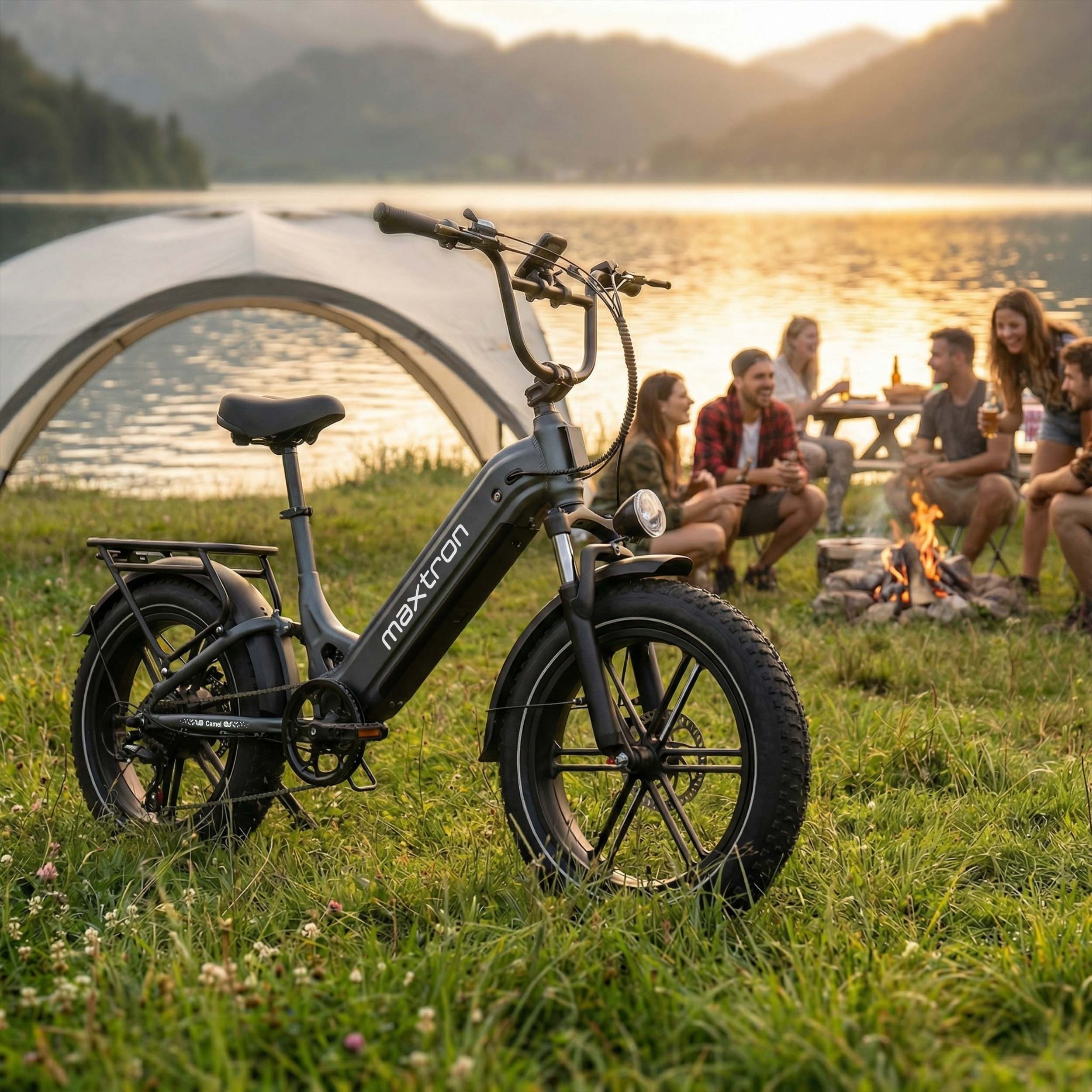 Czarny rower elektryczny na trawie obok jeziora z ludźmi biwakującymi i ogniskiem.