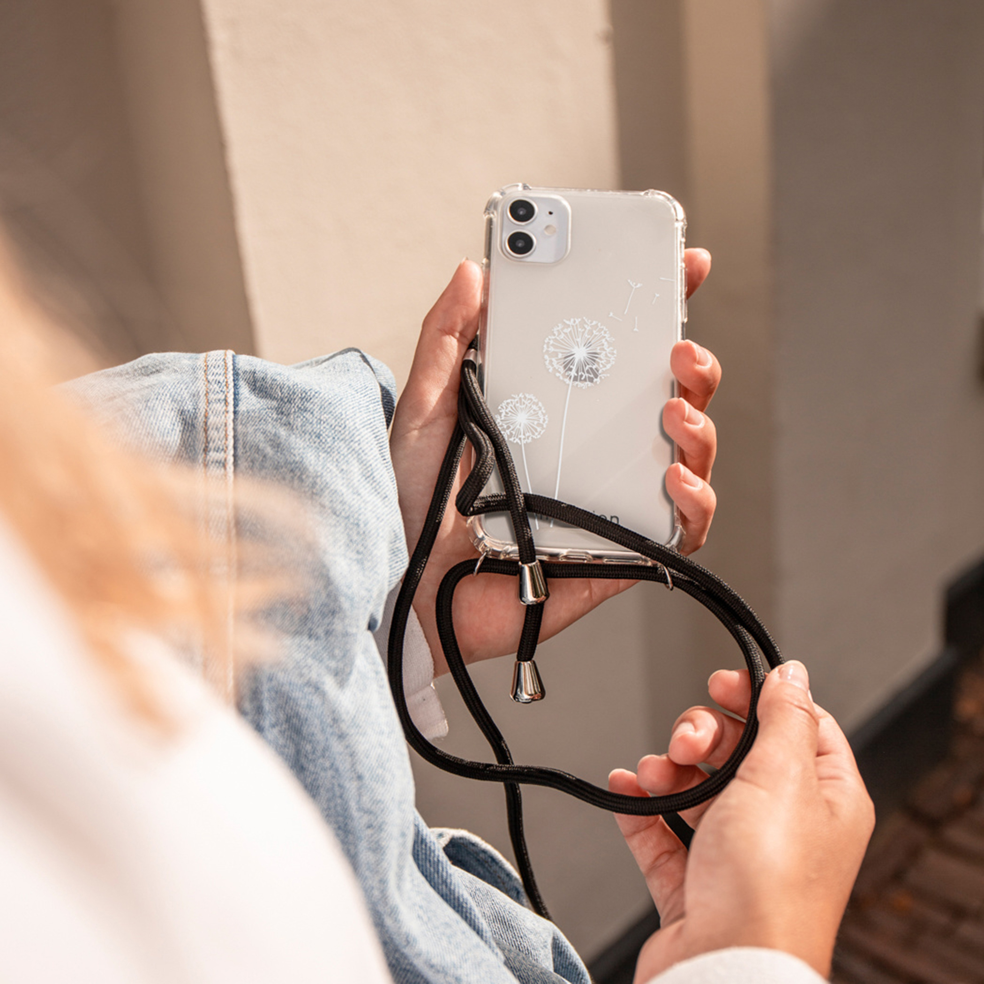 Handen houden een doorzichtige telefoonhoes met paardenbloemprints en een zwarte nekband vast.