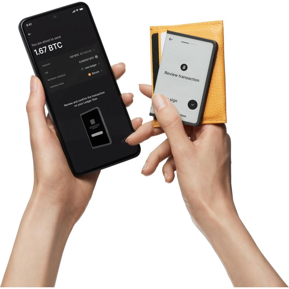 Personne tenant un smartphone avec transaction Bitcoin à côté d'un Ledger Stax dans un étui jaune.