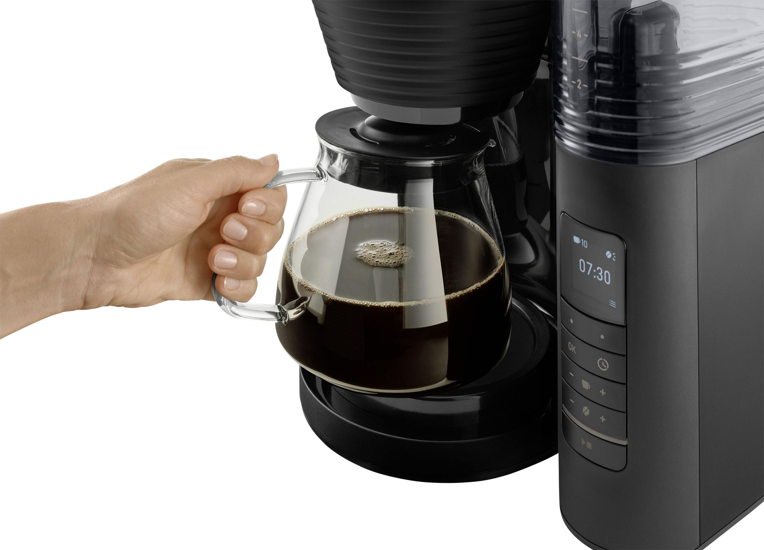 Une main tient une carafe à café en verre à côté d'une cafetière avec écran numérique.