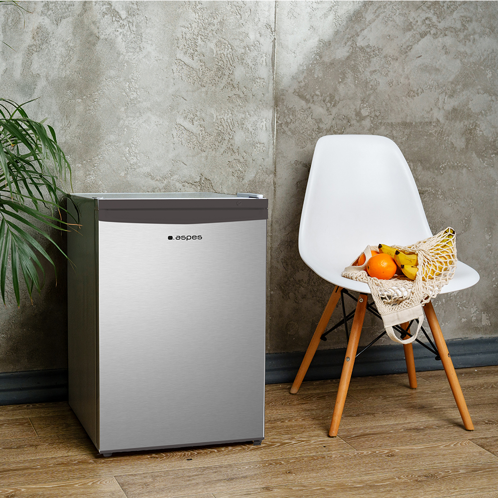 Un refrigerador plateado pequeño junto a una silla blanca con fruta en una bolsa de red.