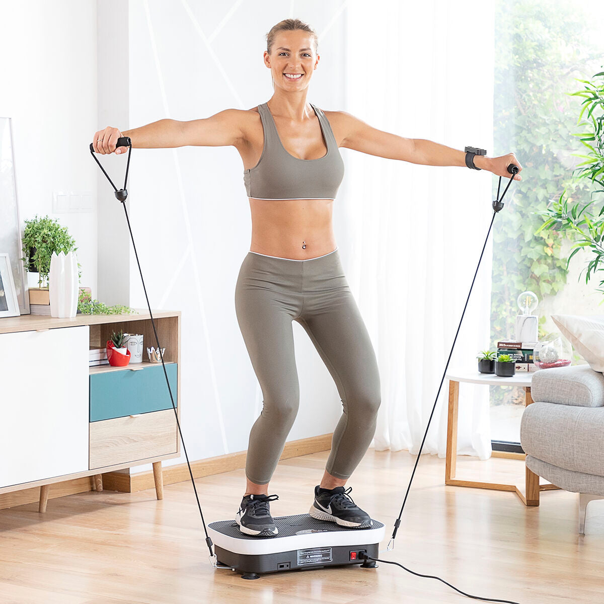 Femme en tenue de sport grise sur plaque vibrante, tenant des bandes de résistance.