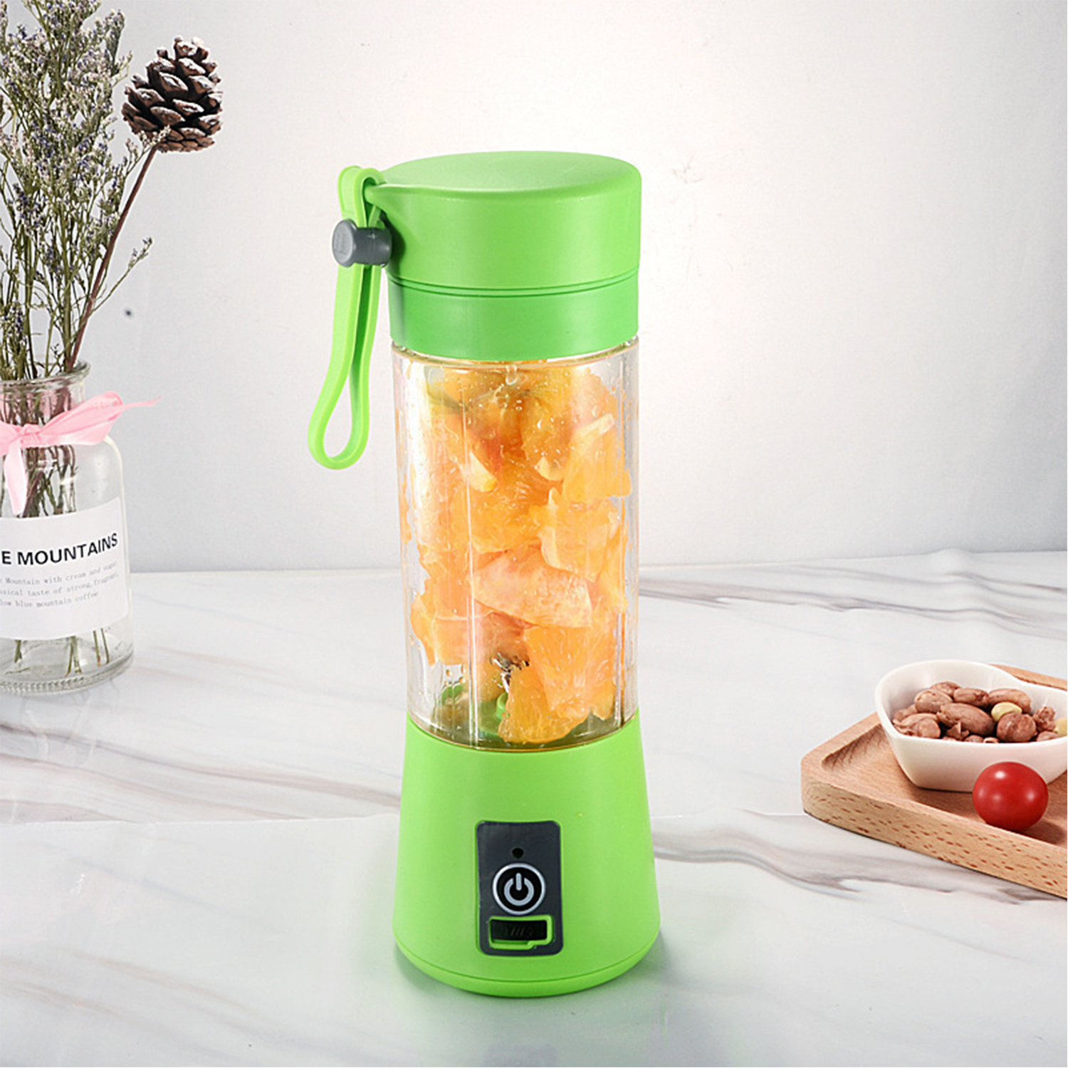 Batidora verde portátil con trozos de naranja, junto a un jarrón y frutos secos.