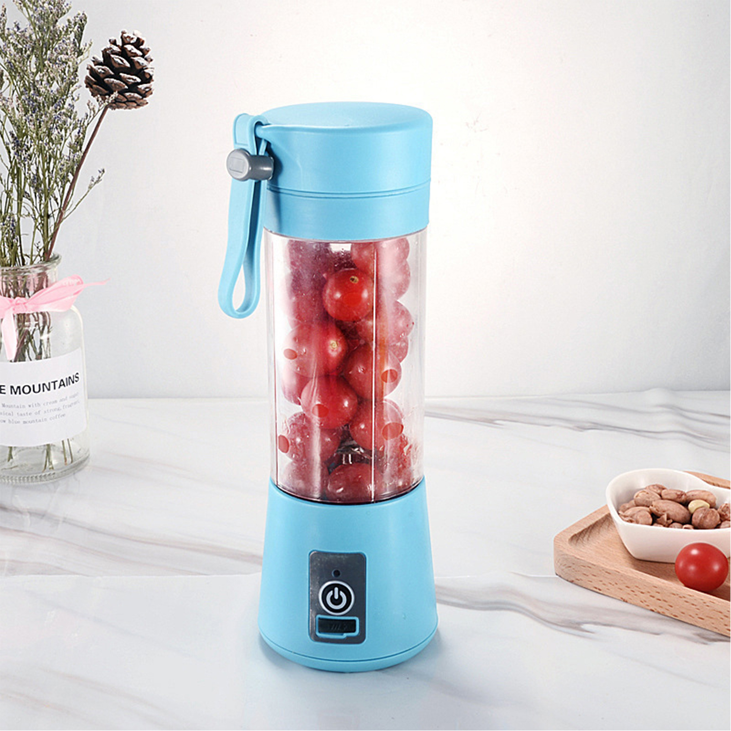 Una licuadora portátil azul llena de bayas rojas se sienta en un mostrador de mármol. Hay frutos secos y un tomate cerca.