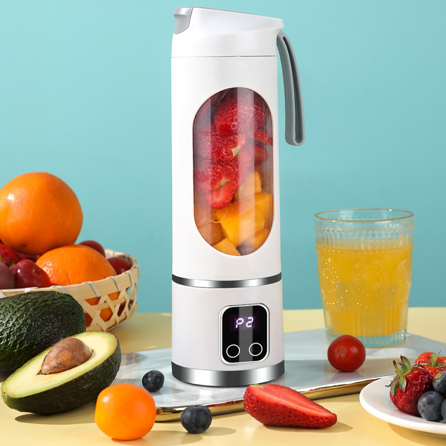 Una batidora portátil blanca llena de fresas y mango, junto a frutas y un vaso de zumo.