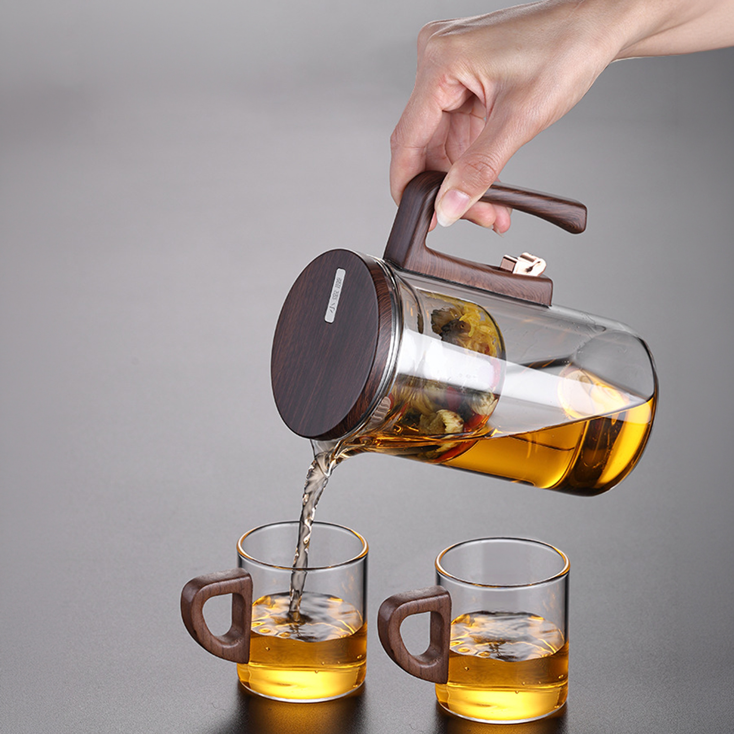 Una mano vierte té dorado de una jarra de vidrio con asas de madera en dos tazas.