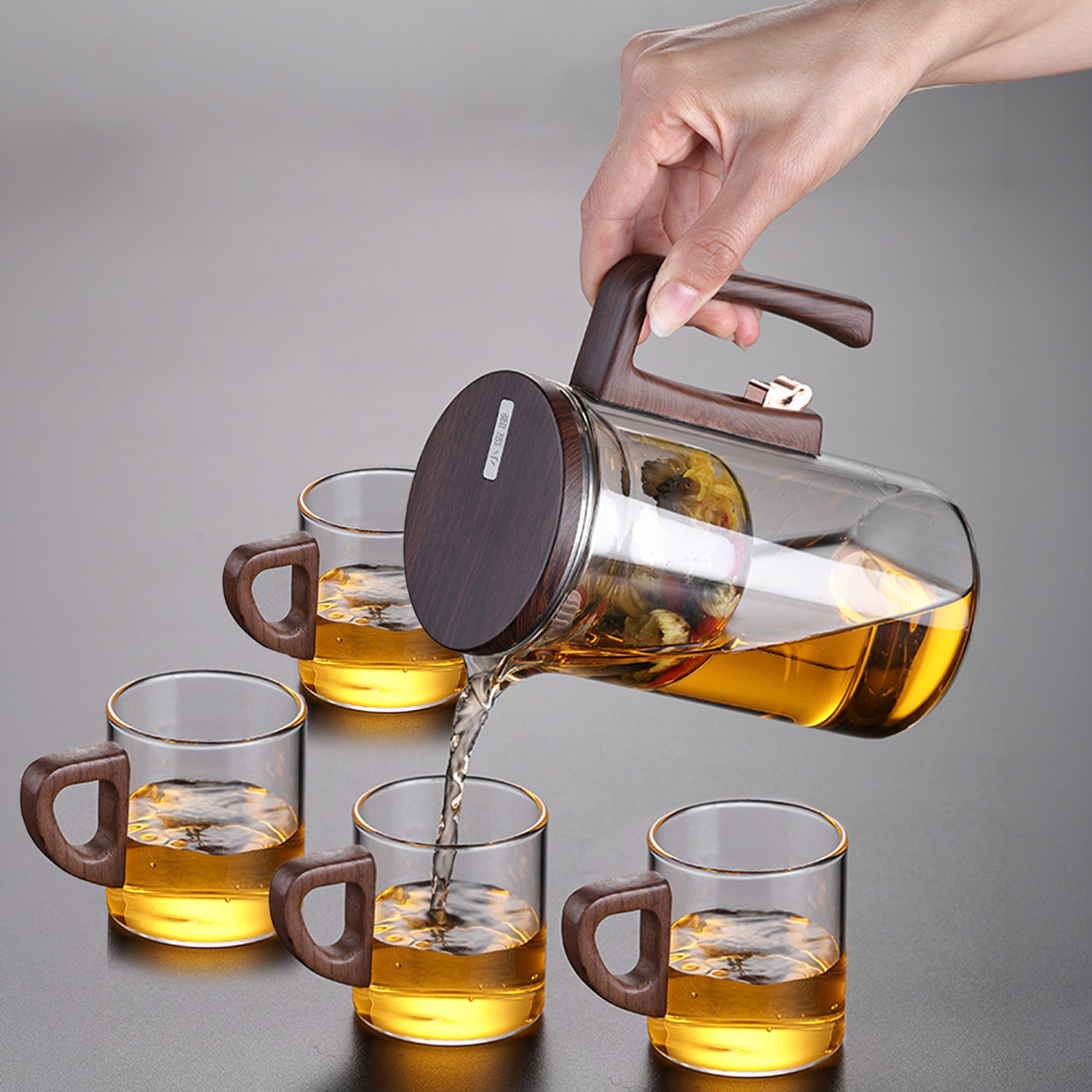 Una mano vierte té de una tetera de cristal con asa de madera en cuatro tazas pequeñas.
