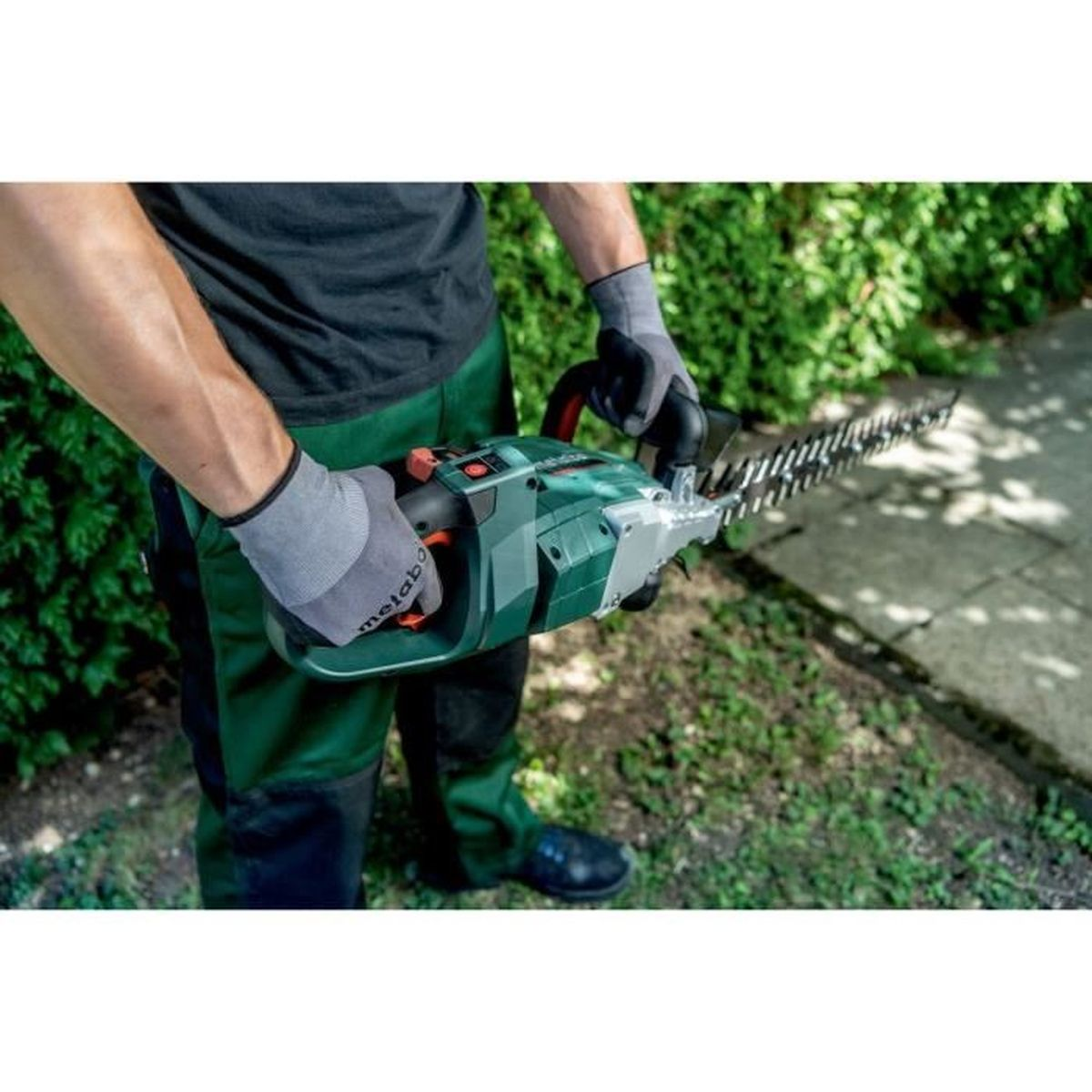 Mann mit grüner Heckenschere im Garten, trägt Handschuhe und Arbeitskleidung.