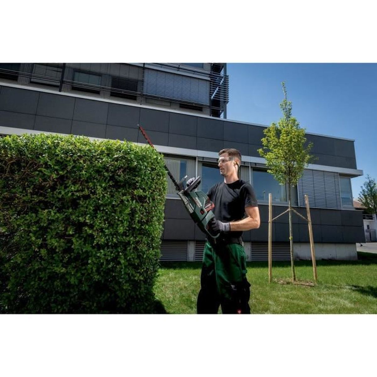 Mann trimmt Hecke mit Heckenschere vor modernem Gebäude an einem sonnigen Tag.