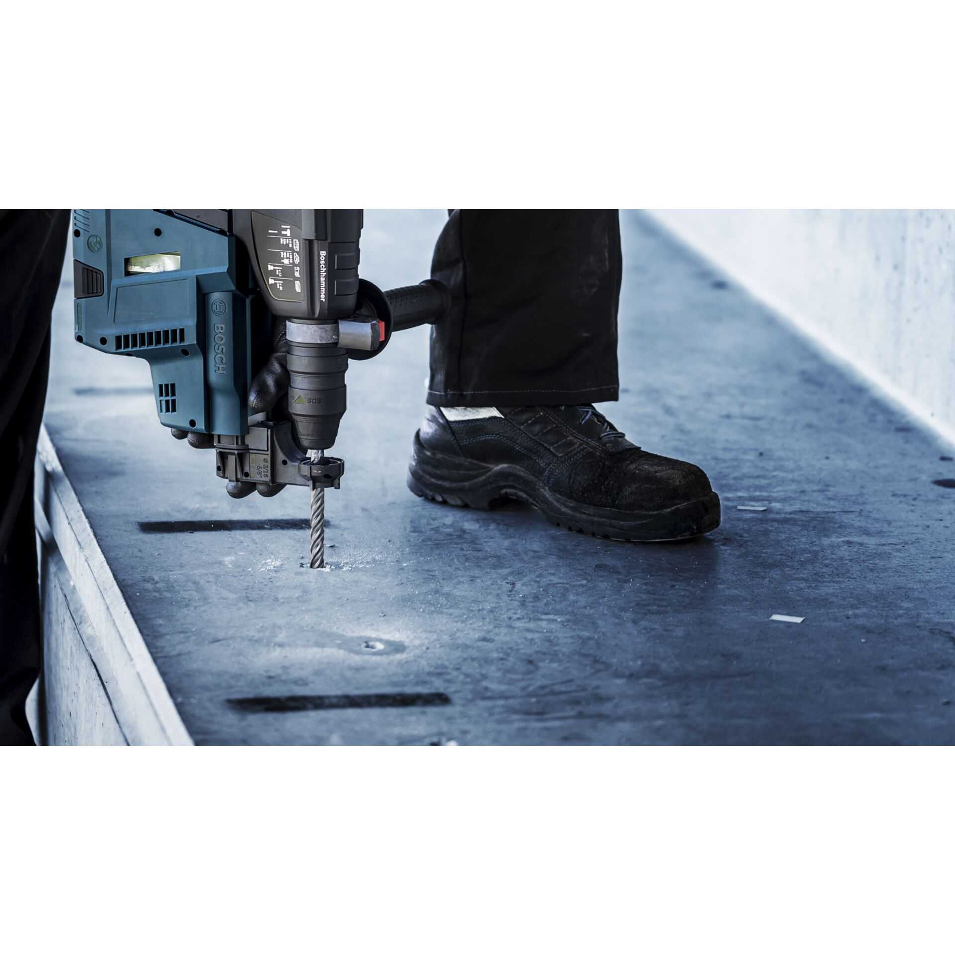Mann bohrt mit Bosch Hammerbohrer in Beton. Schwarzer Schuh und Hose sichtbar.