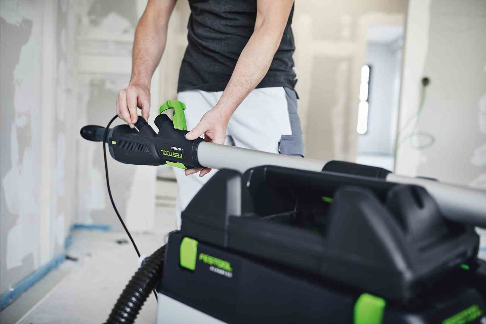 Ein Mann hält ein Festool Schleifgerät mit grünem Griff, verbunden mit einem Staubsauger.