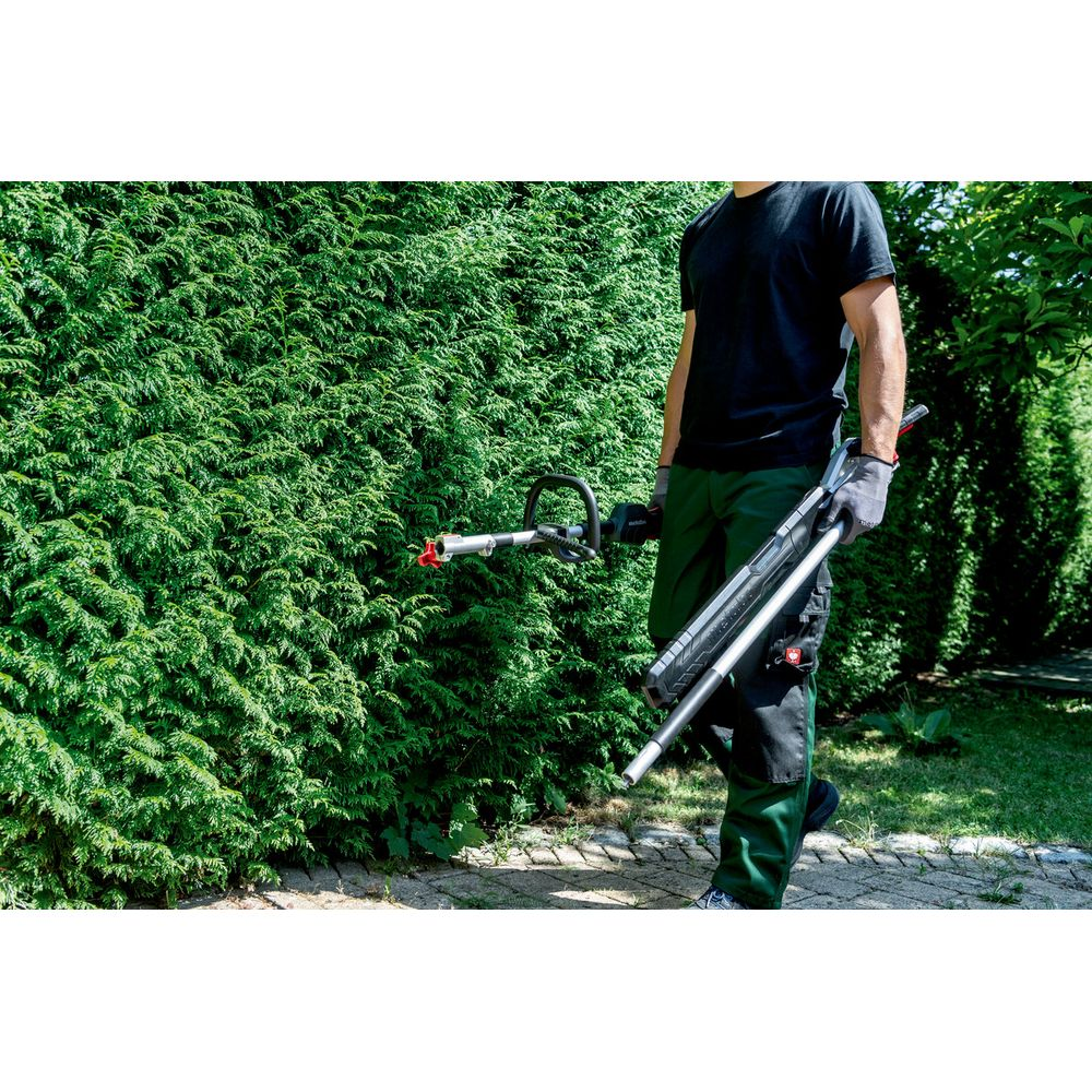 Mann in schwarzem Hemd und grüner Hose hält Heckenschere vor grüner Hecke.