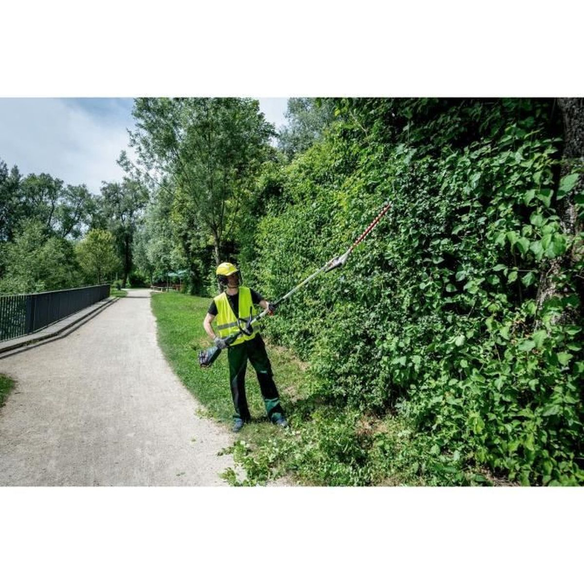 Mann mit Heckenschere auf einem Feldweg neben einer dichten Hecke.