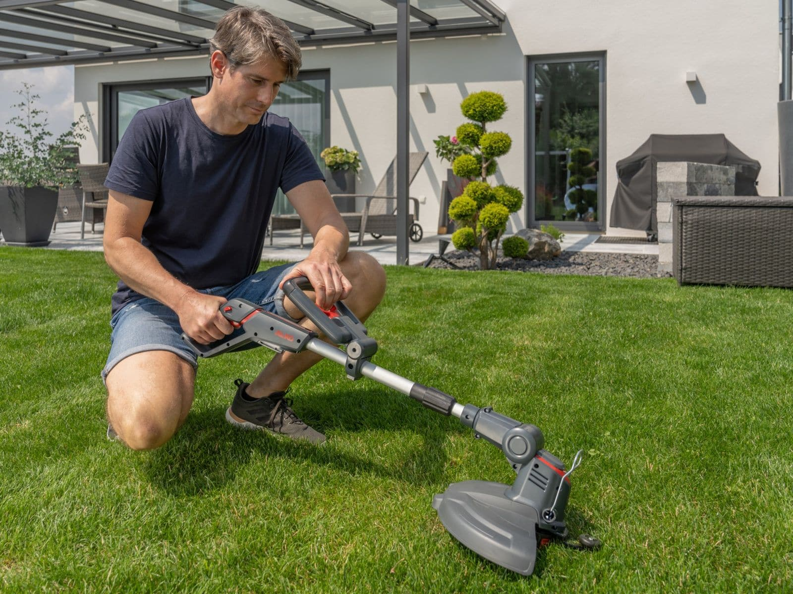 Mann kniet im Garten und hält einen Rasentrimmer. Haus und Sträucher im Hintergrund.