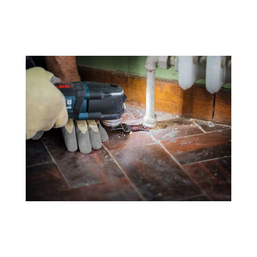 Eine Hand mit Handschuhen benutzt ein oszillierendes Werkzeug auf einem Holzboden neben einem Heizkörperrohr.