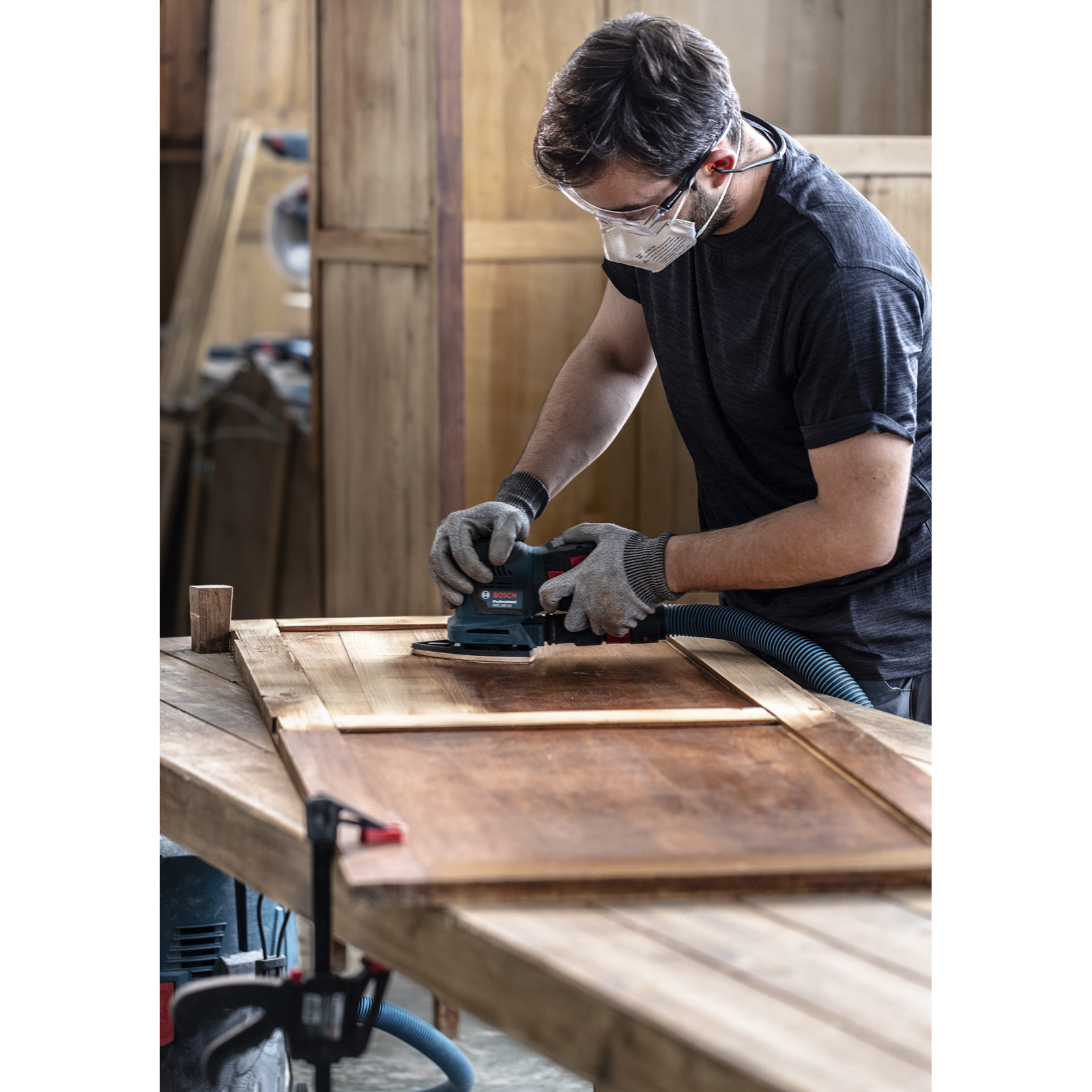 Mann schleift Holz mit Exzenterschleifer, trägt Maske und Schutzbrille.