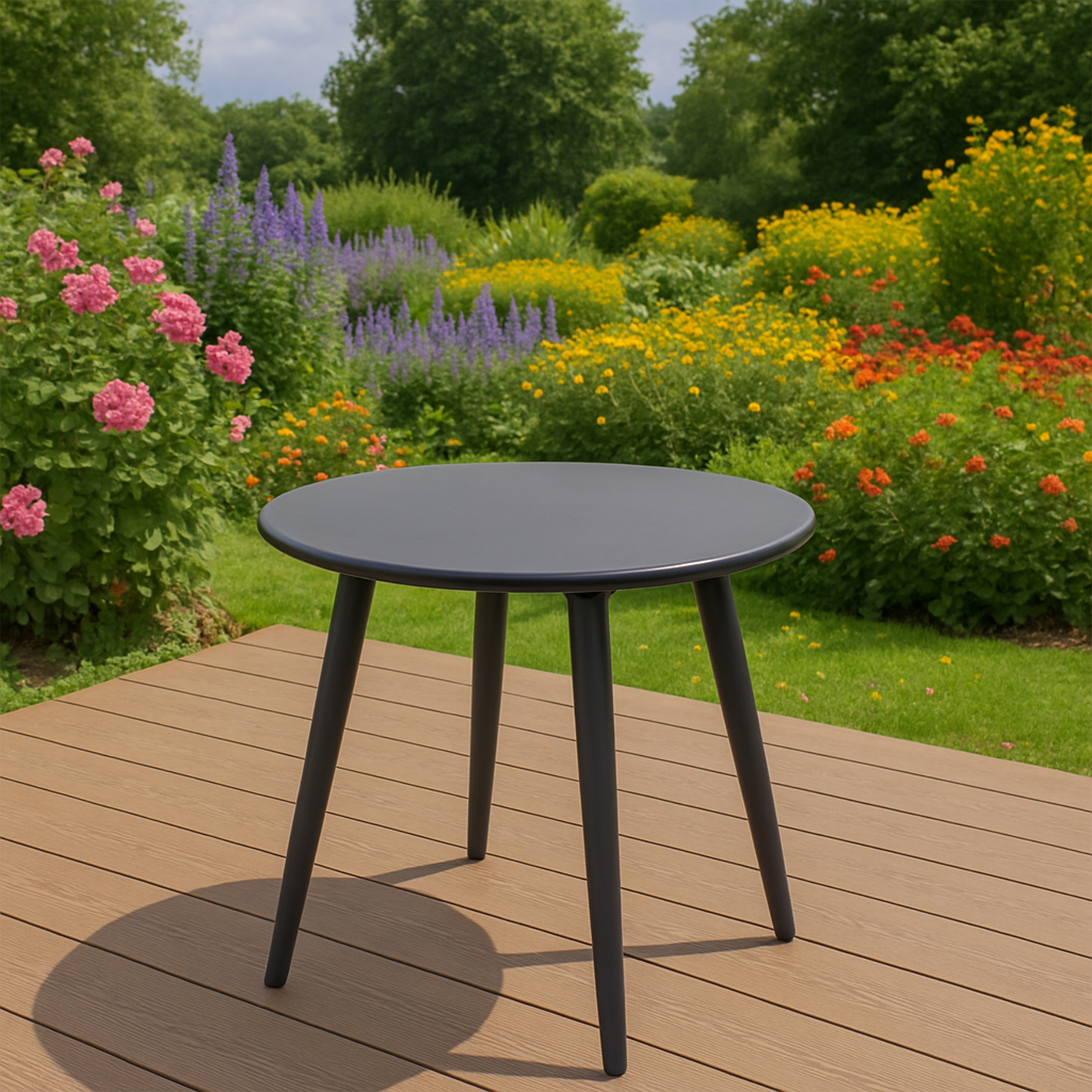 Ein runder, dunkler Tisch steht auf einer Holzterrasse vor einem blühenden Garten.
