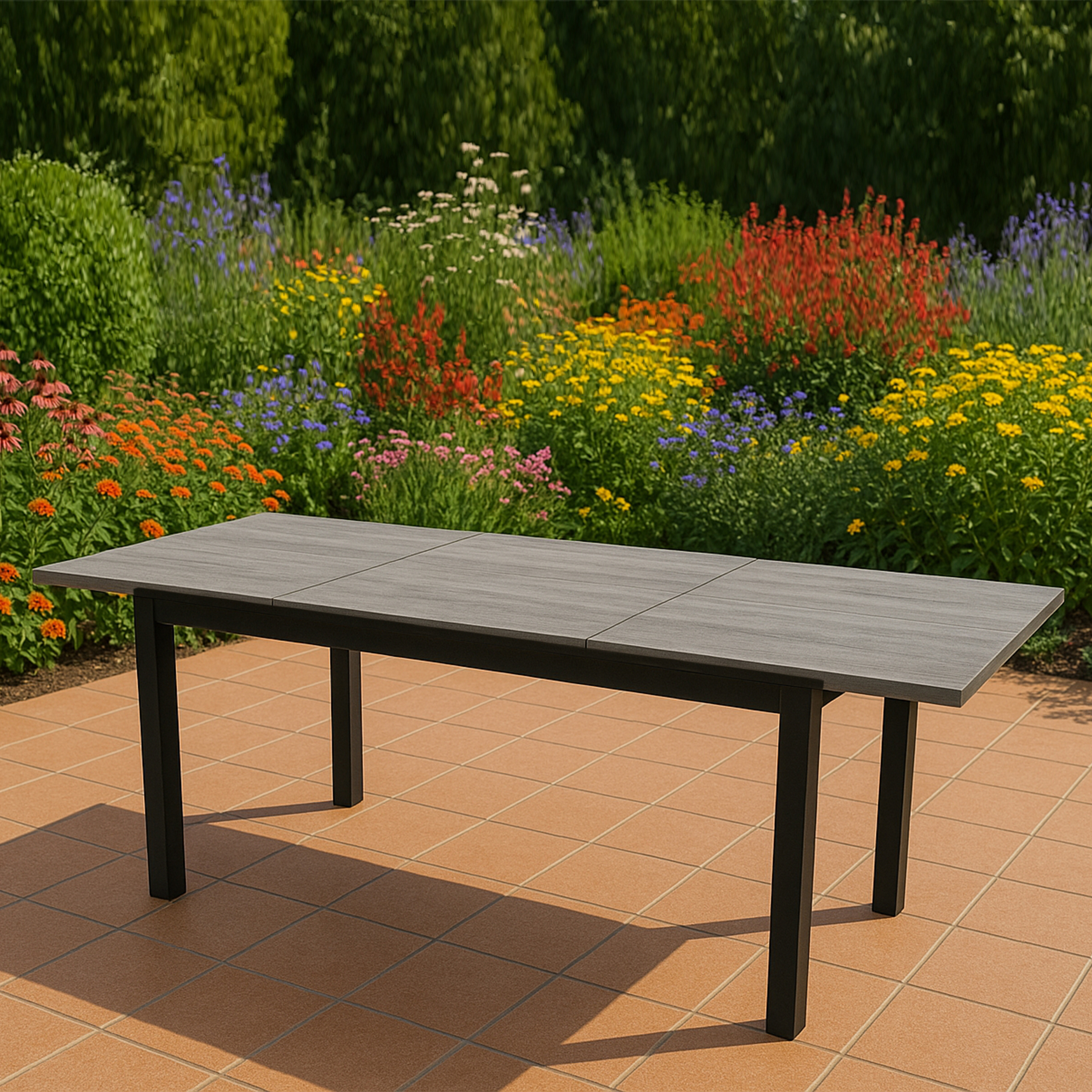 Ein grauer Tisch steht auf einer Terrakotta-Terrasse vor einem bunten Blumenbeet.