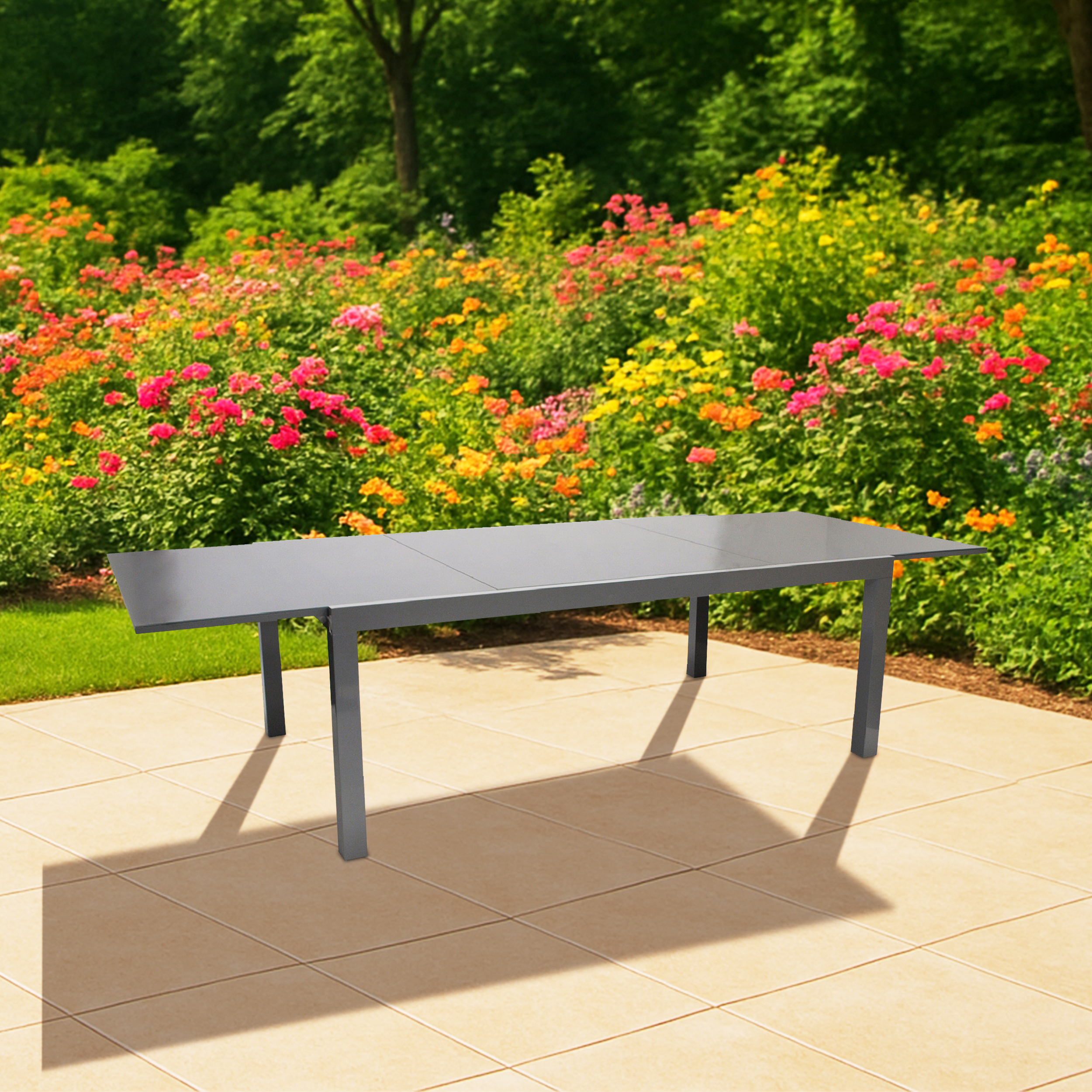 Ein grauer Tisch steht auf einer gefliesten Terrasse vor blühenden Blumen und grünen Bäumen.