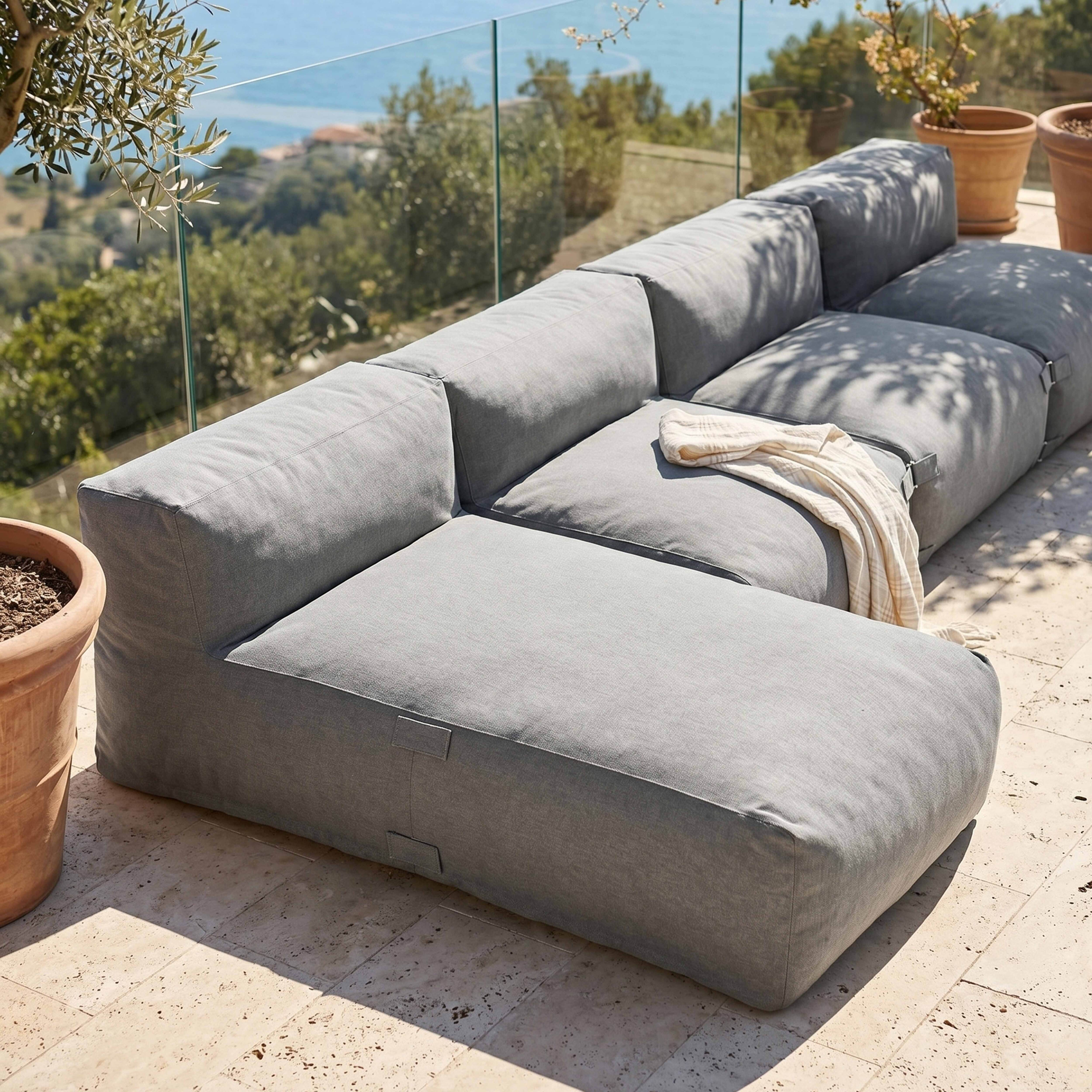 Graues Modulsofa auf einer Steinterrasse mit Meerblick und Bäumen.