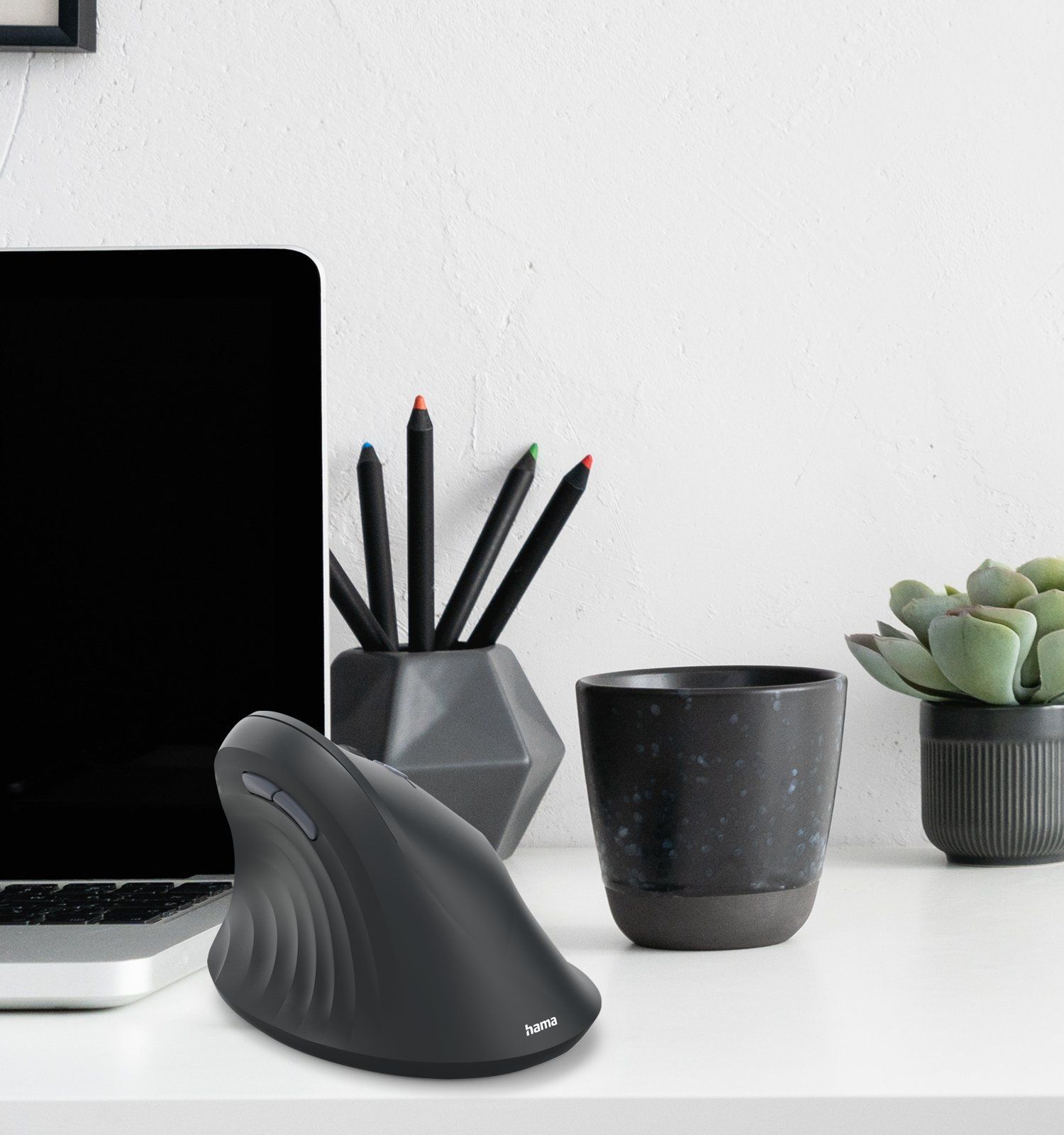 Eine schwarze ergonomische Maus steht auf einem weißen Schreibtisch neben einem Laptop, Stiften und einer Pflanze.